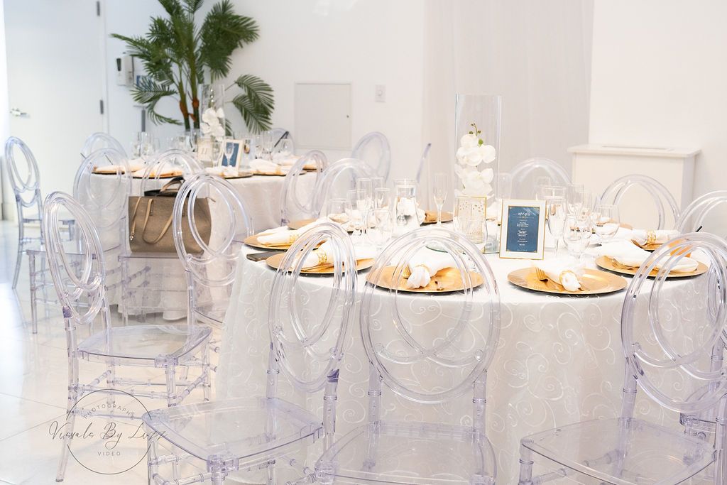 Modern wedding reception setup with ghost chairs and gold charger plates in an elegant white venue.