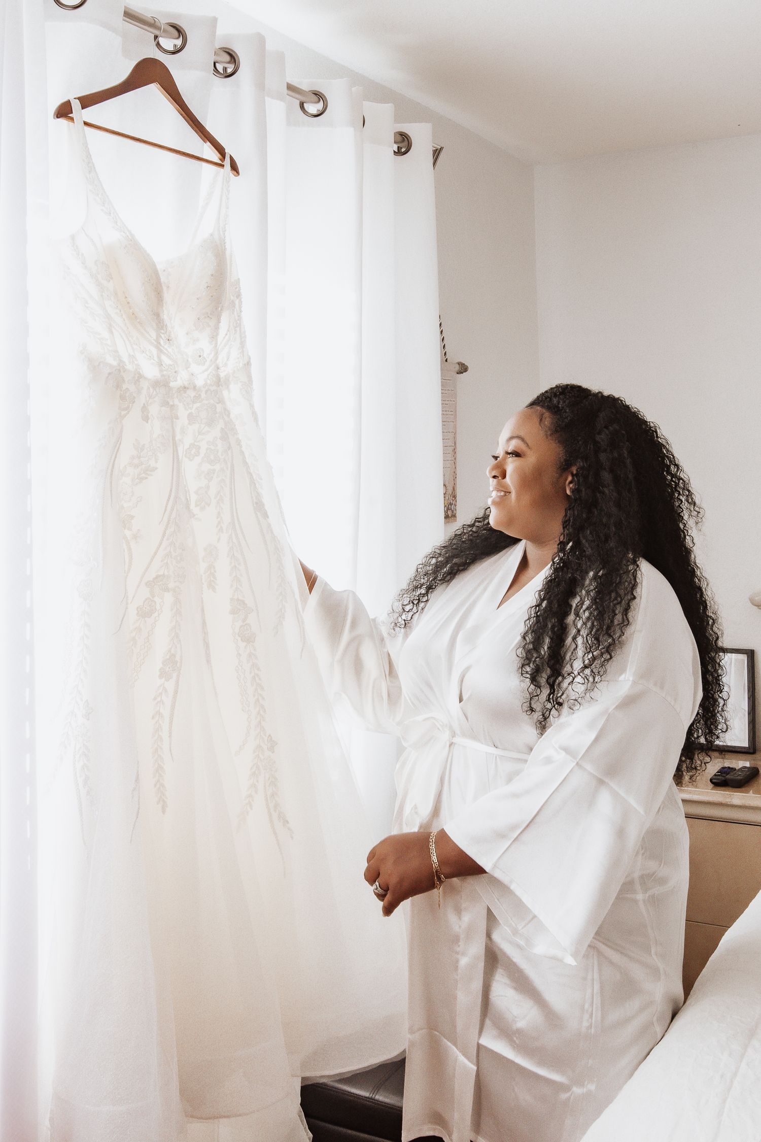White wedding dress hangs on display in bridal suite with natural lighting.