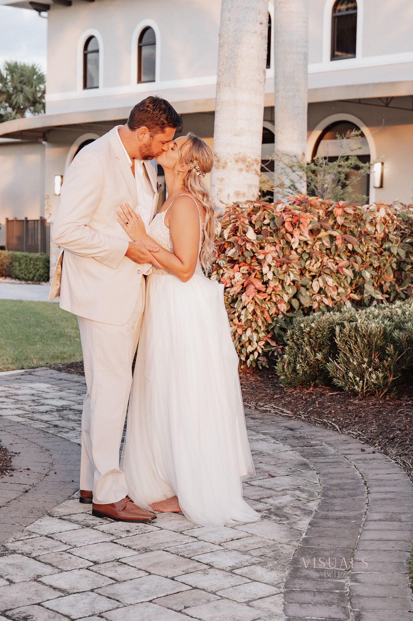A romantic wedding couple shares a tender moment on a stone pathway beside white columns and lush foliage.
