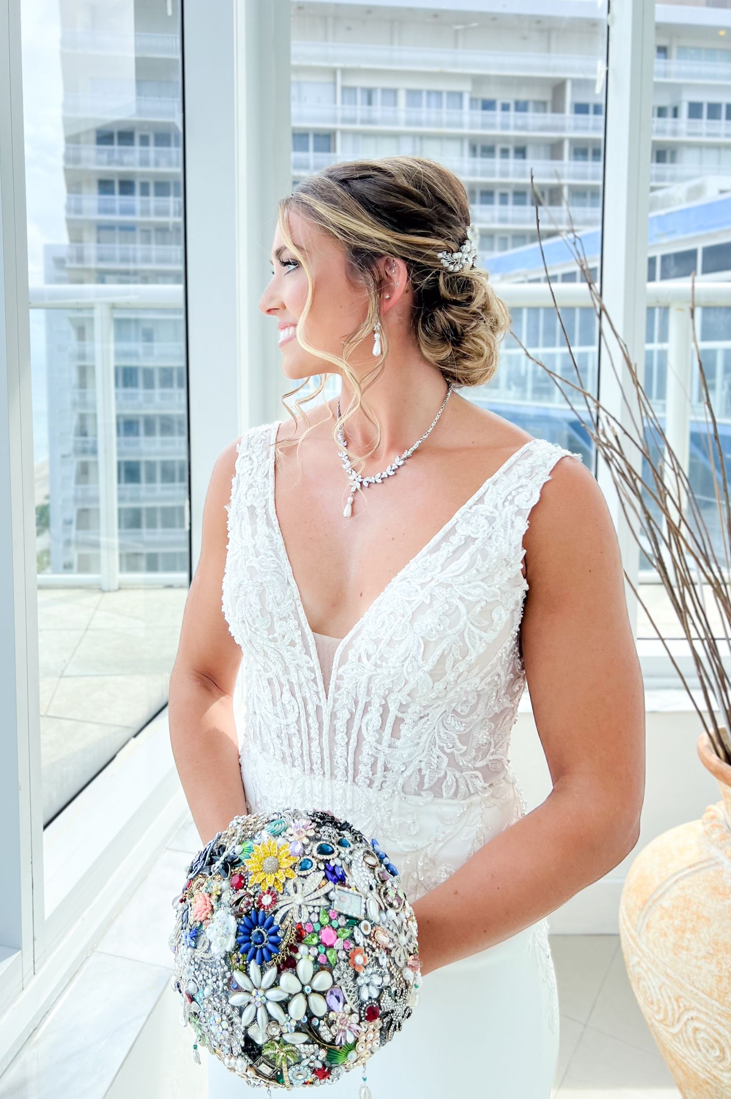 Elegant bride in white lace dress holding sparkly brooch bouquet near window.