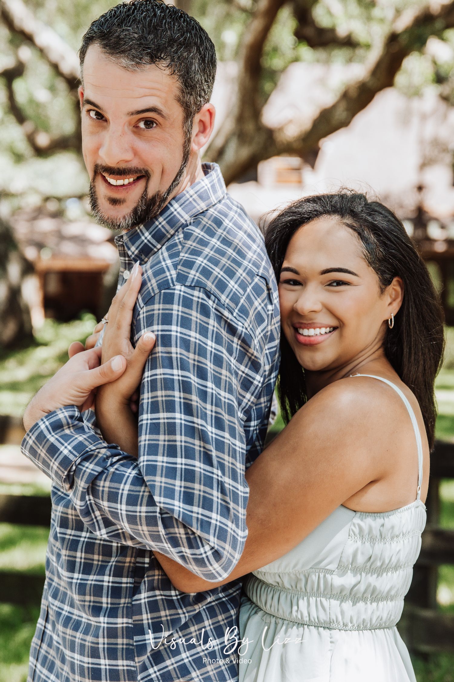 A joyful couple embraces outdoors in a garden setting, with a plaid shirt and white dress against natural greenery.