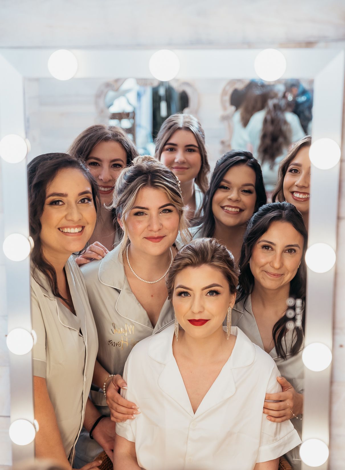 Bridal party in matching white robes posing together in dressing room with illuminated vanity mirror.