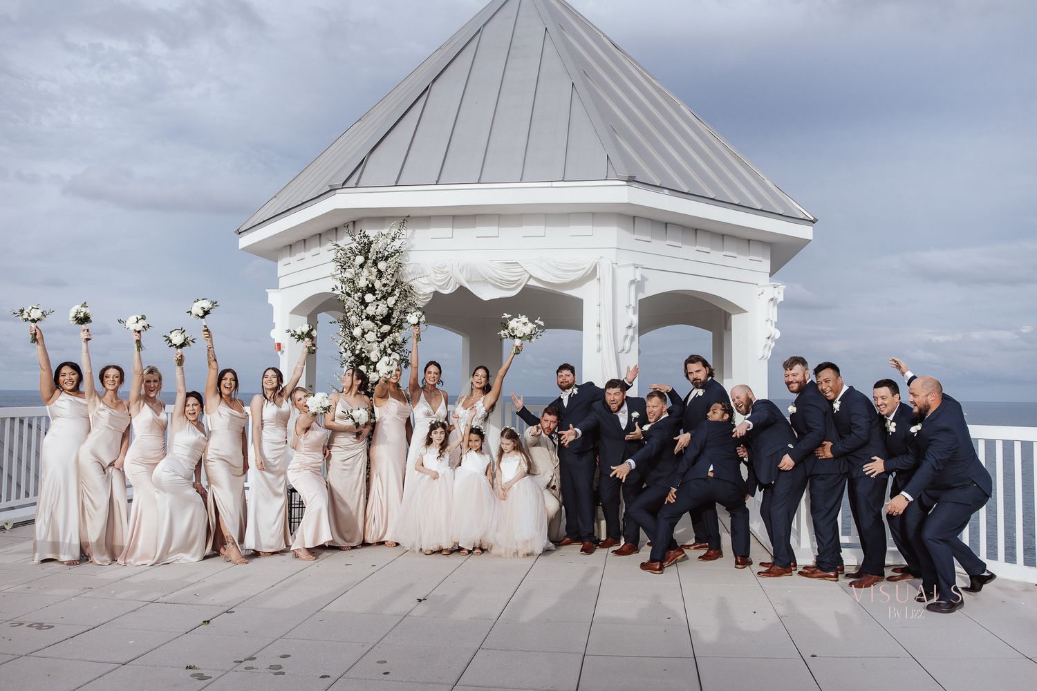 A wedding party poses together under a white gazebo on a waterfront deck with their arms raised in celebration.