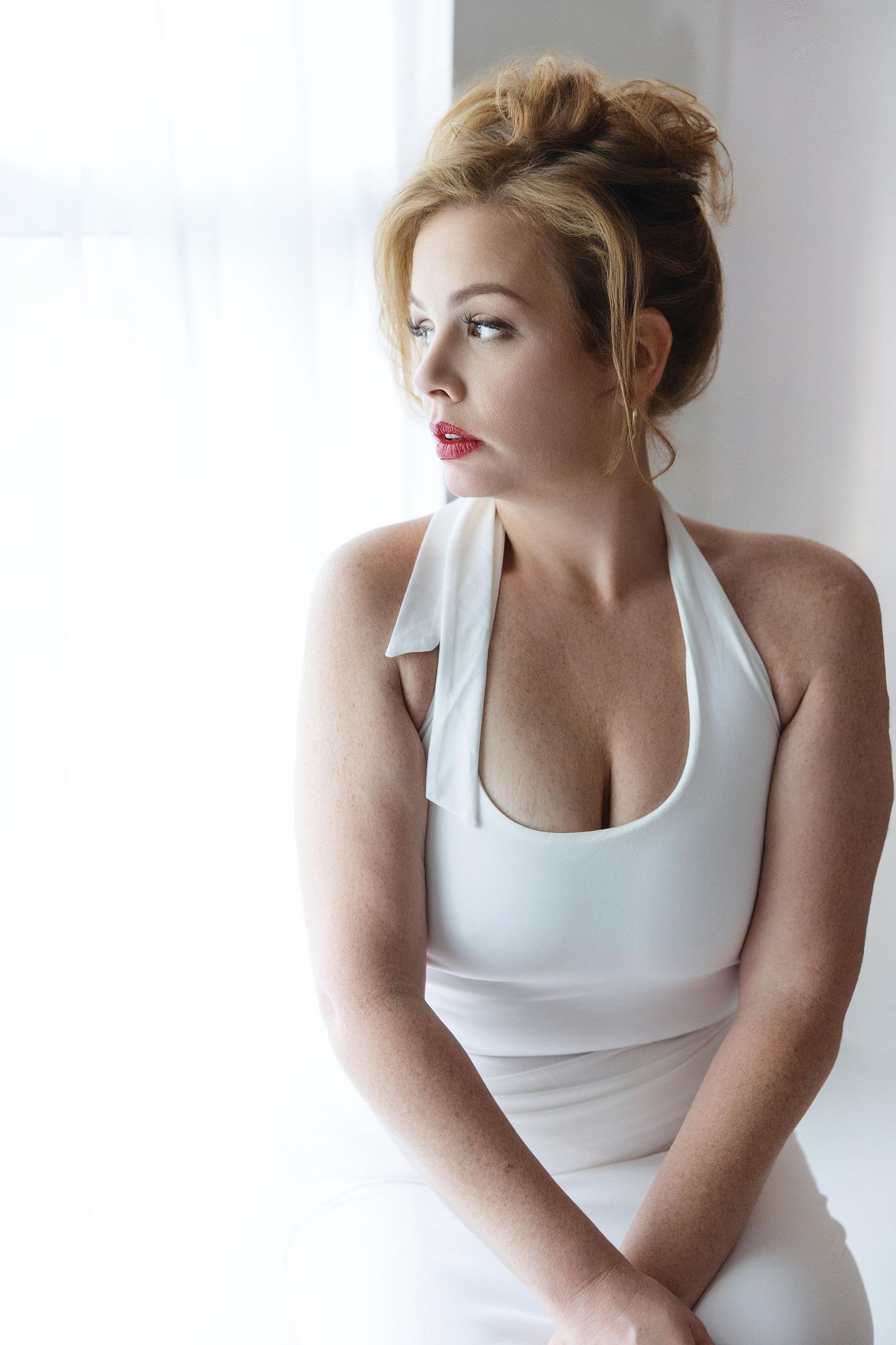 Elegant blonde woman in white halter dress sitting by bright window with red lips and updo hairstyle.