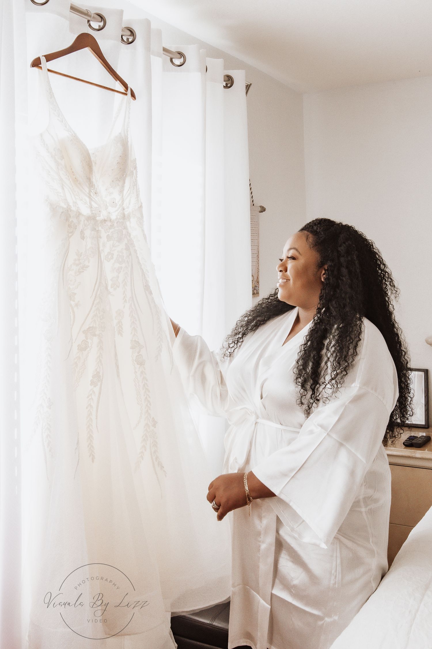 A joyful woman in a white robe stands near hanging wedding dresses while getting ready.