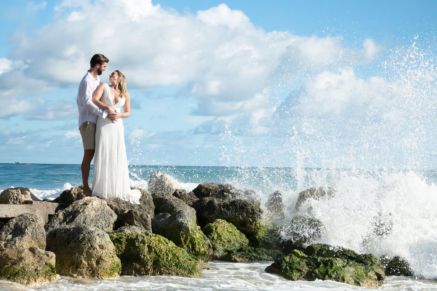 Couple in white attire poses on rocky beach shore as ocean waves crash dramatically against the rocks in the background.