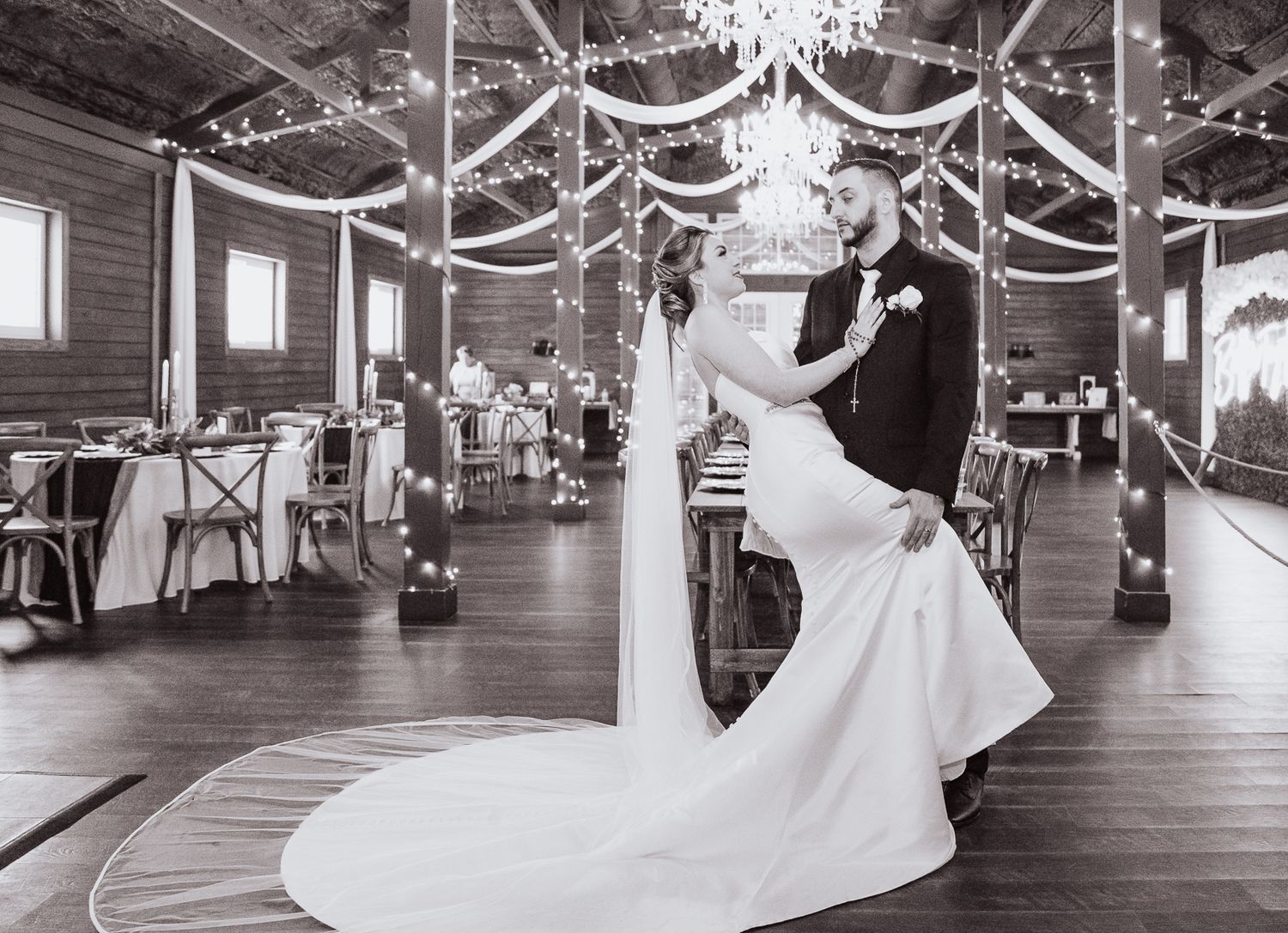 Black and white photo of newlywed couple dancing in elegant ballroom under crystal chandeliers.