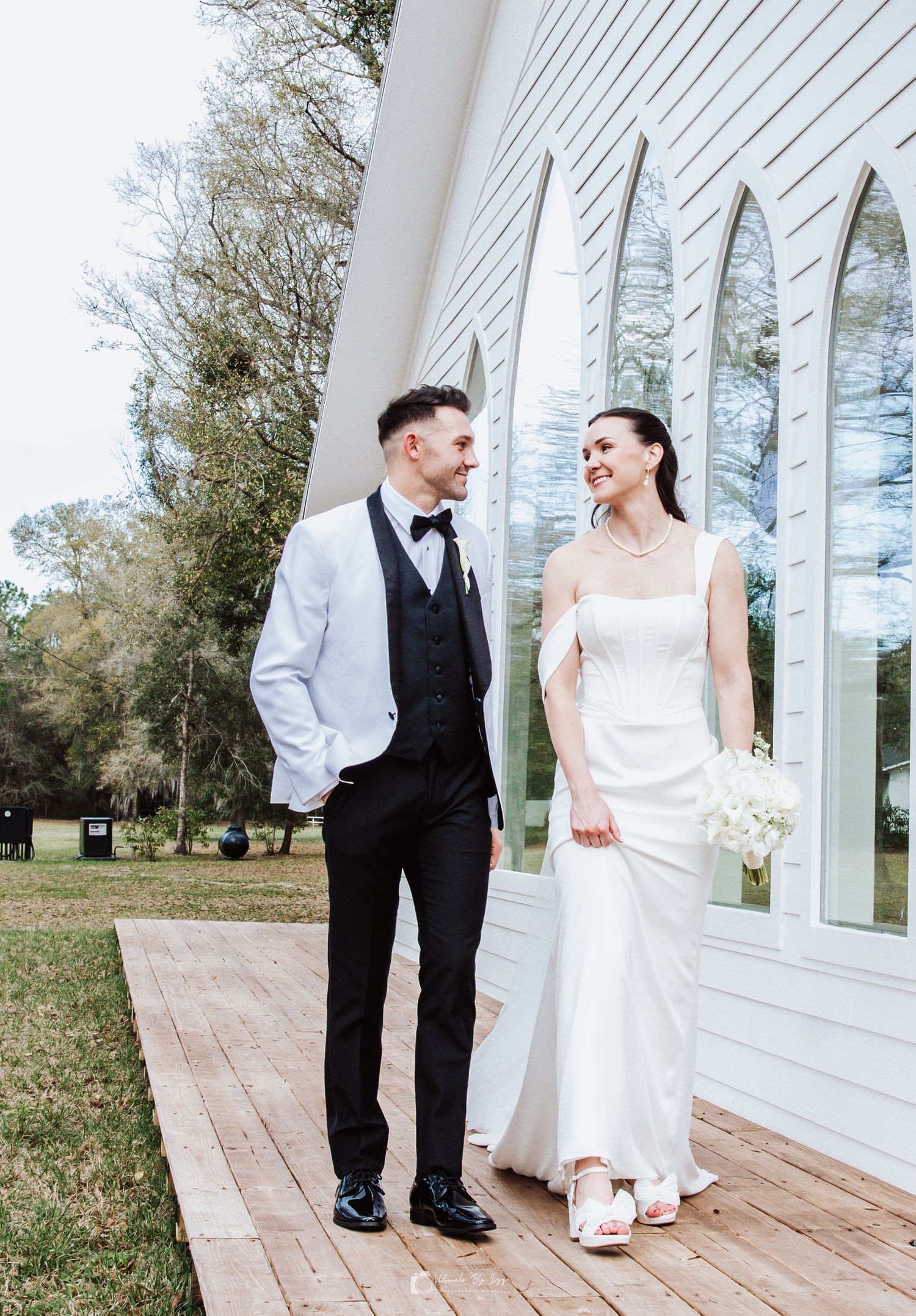 A couple in wedding attire stands on a wooden deck beside a white church with tall reflective spires.