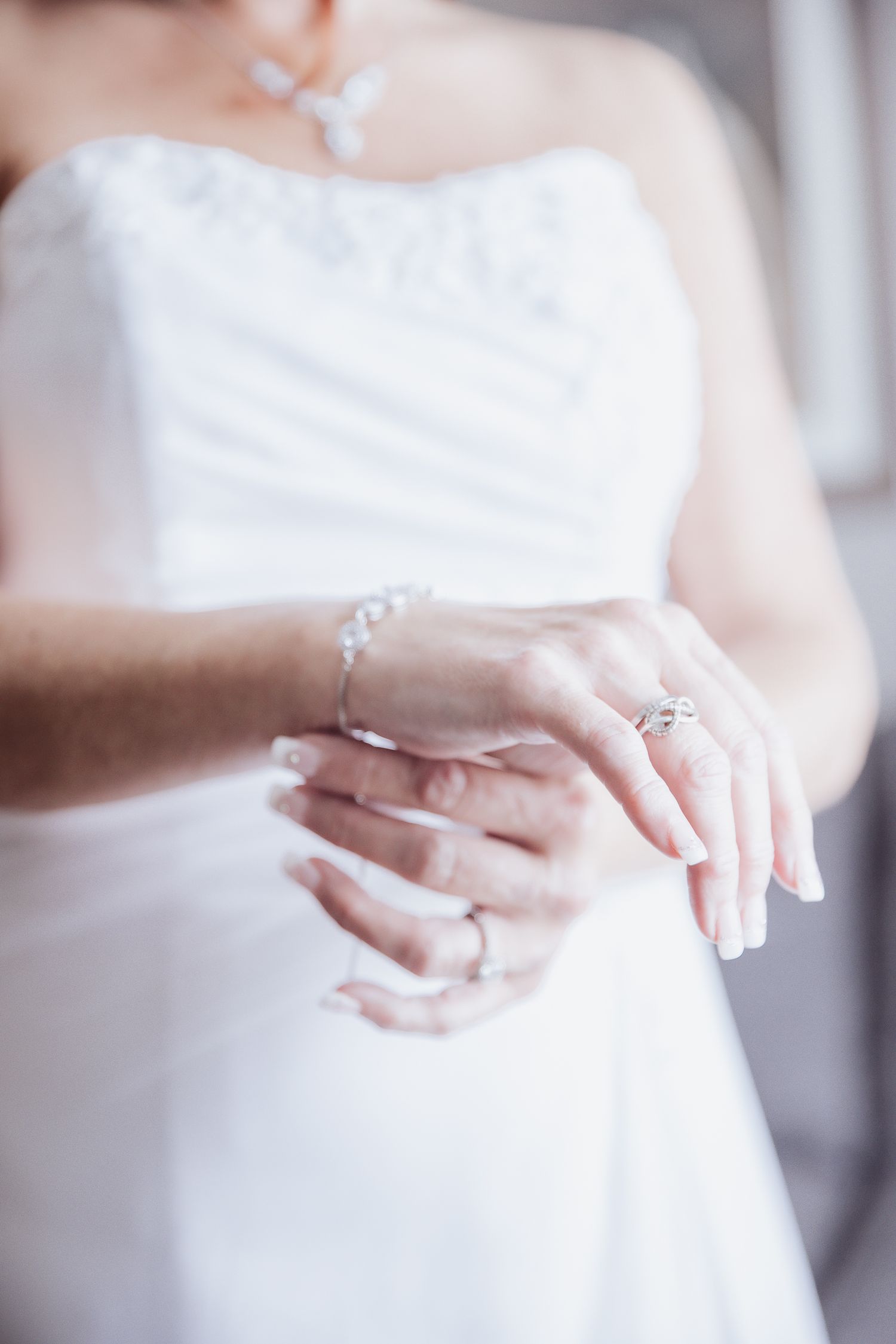 Close-up of delicate bridal jewelry and hands against a white wedding dress.