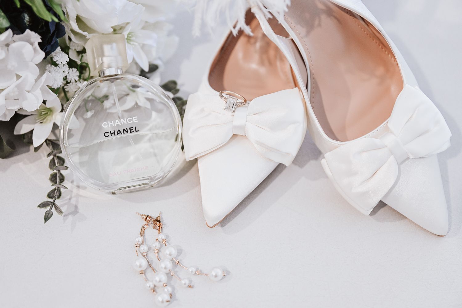 Elegant white bridal shoes with bows next to Chanel perfume bottle and white floral arrangement on light surface.
