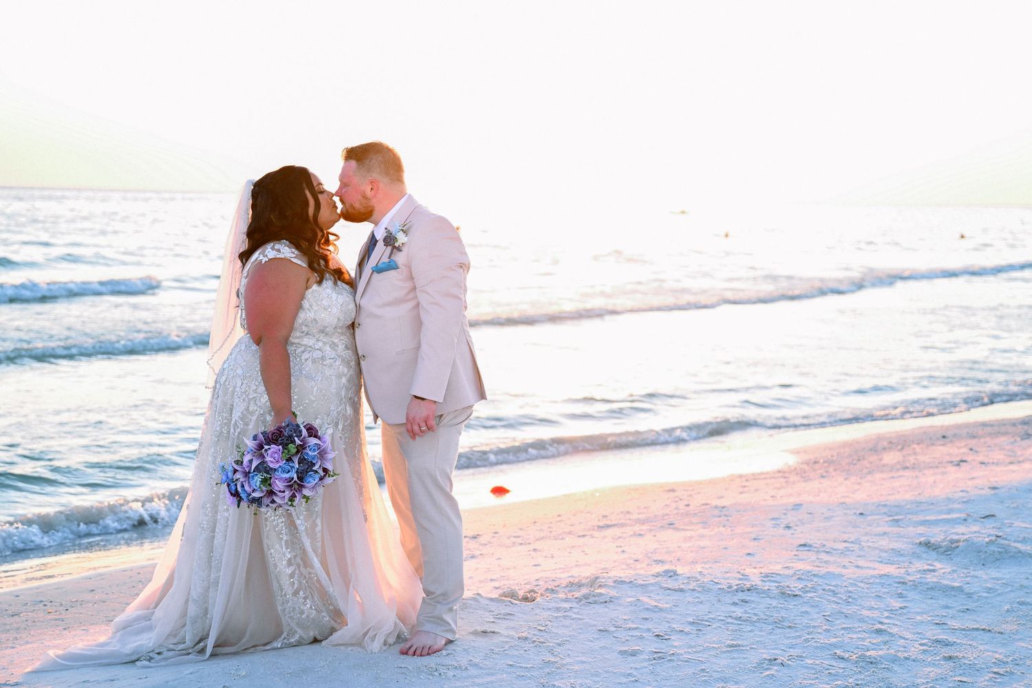 Bride and groom share a romantic kiss on a sandy beach at sunset, bride holding a colorful bouquet.
