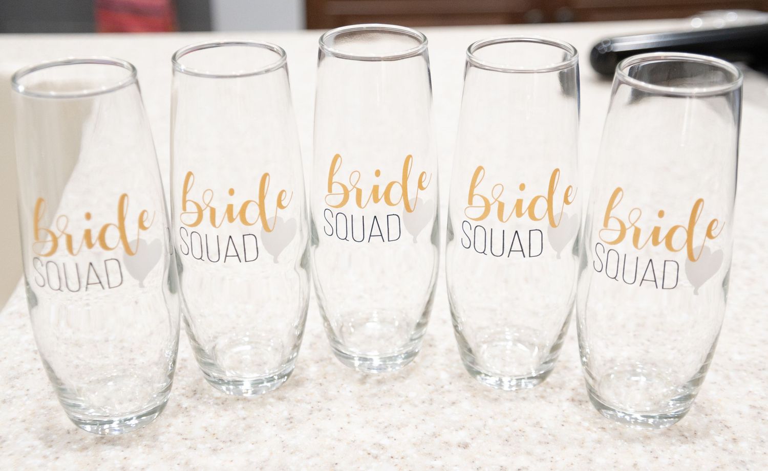 Five champagne flutes with gold 'bride squad' lettering arranged on white surface.