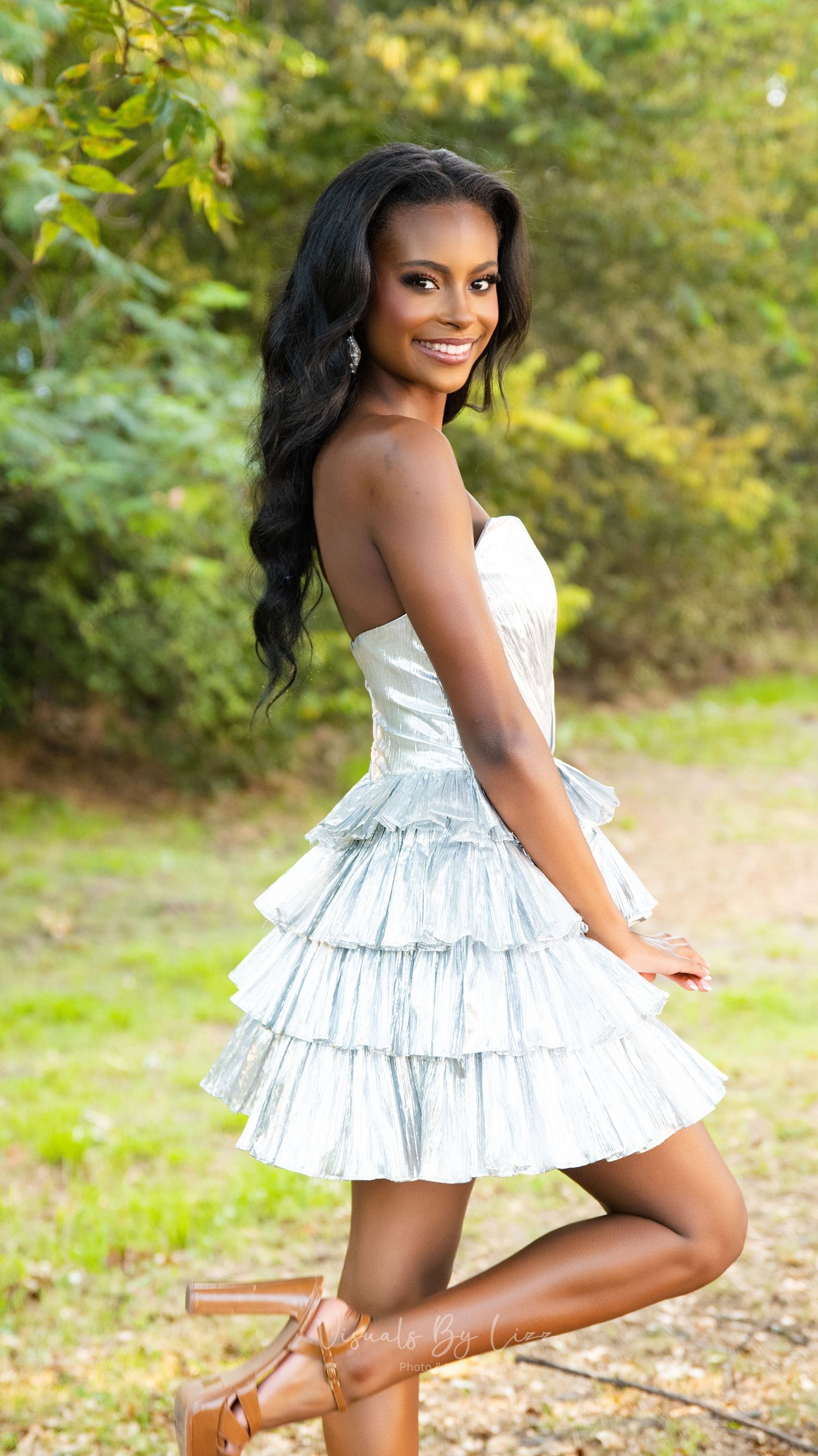 A person poses outdoors in a white ruffled strapless dress with tiered skirt and brown sandals against a natural backdrop.