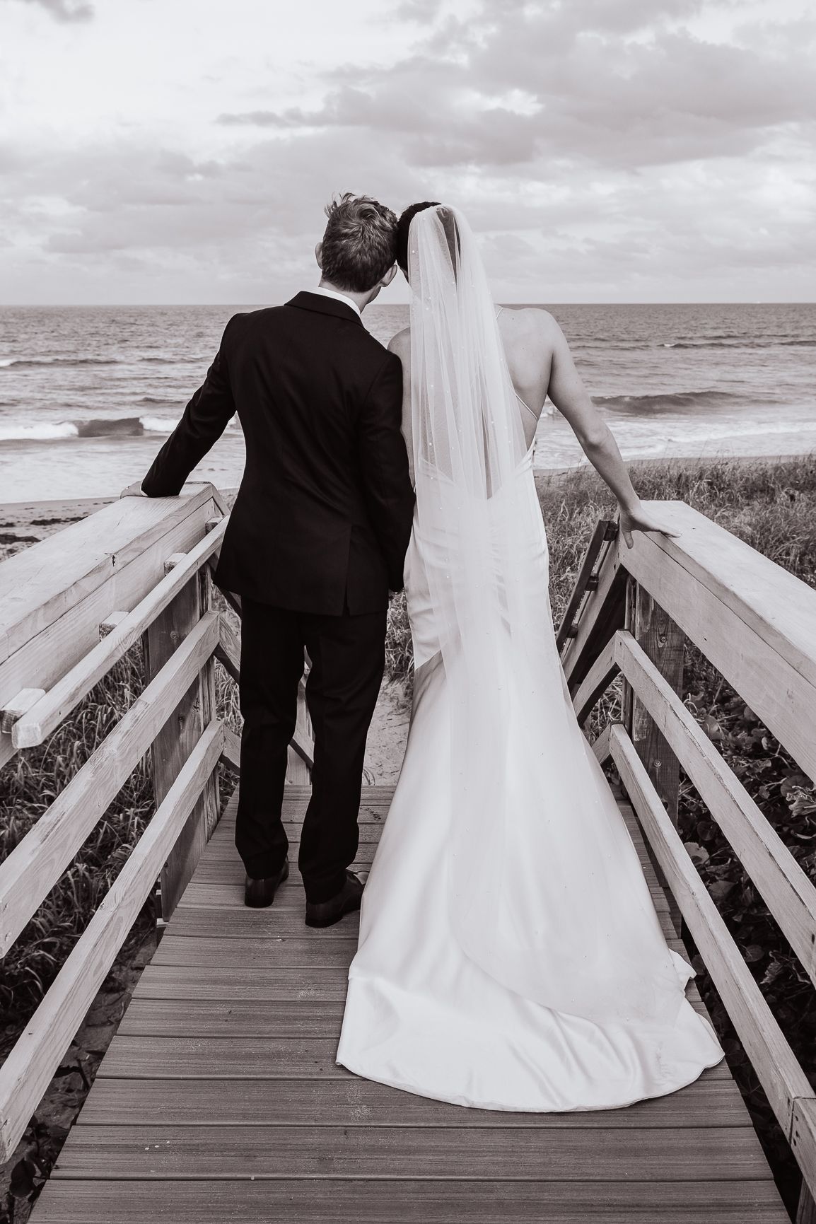A black and white series of wedding photos showing a couple walking away on a wooden boardwalk by the ocean.