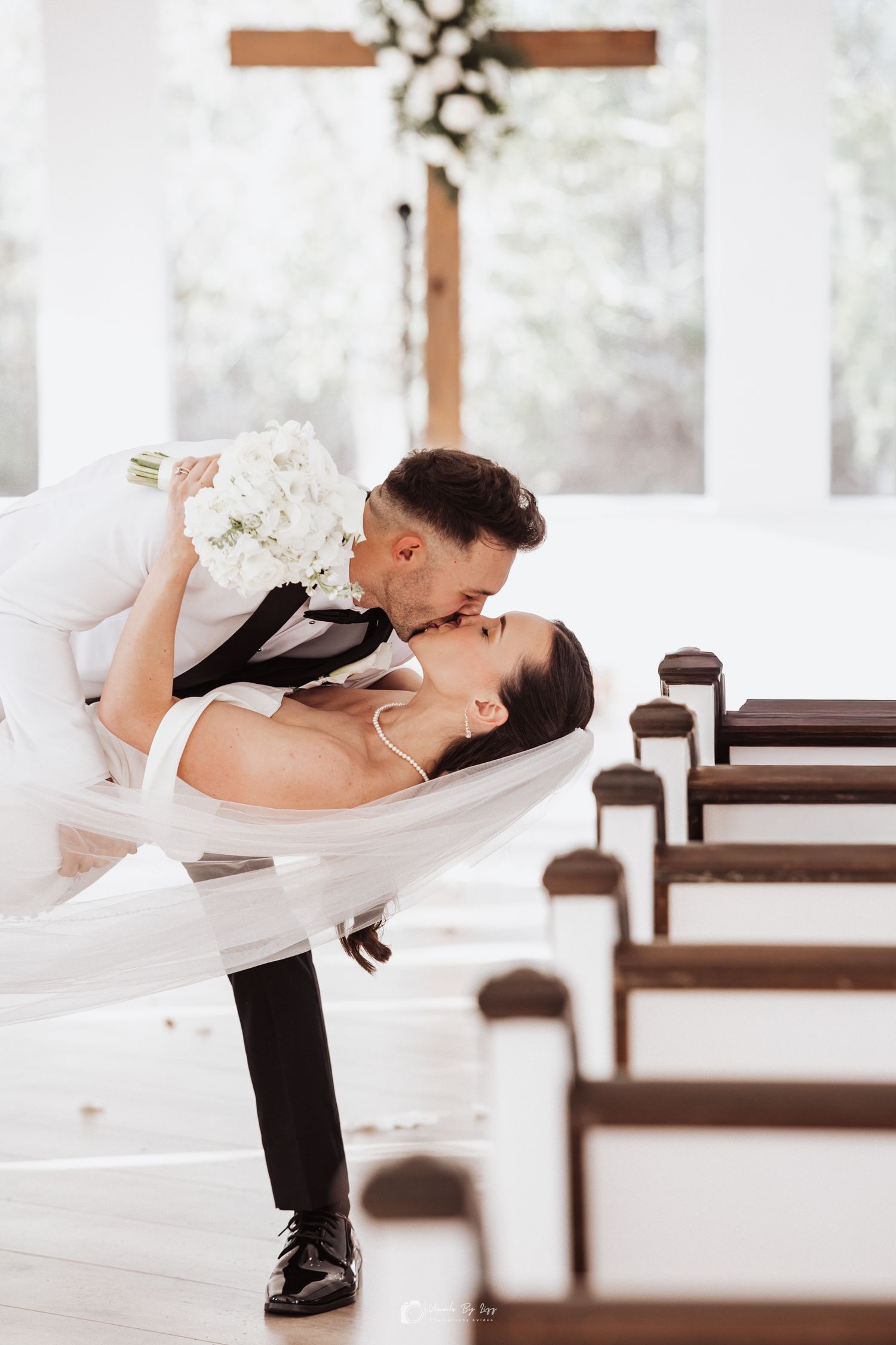 A romantic wedding couple dips in a dramatic pose inside a bright white chapel with wooden pews. The Whitewood Ranch, Fl