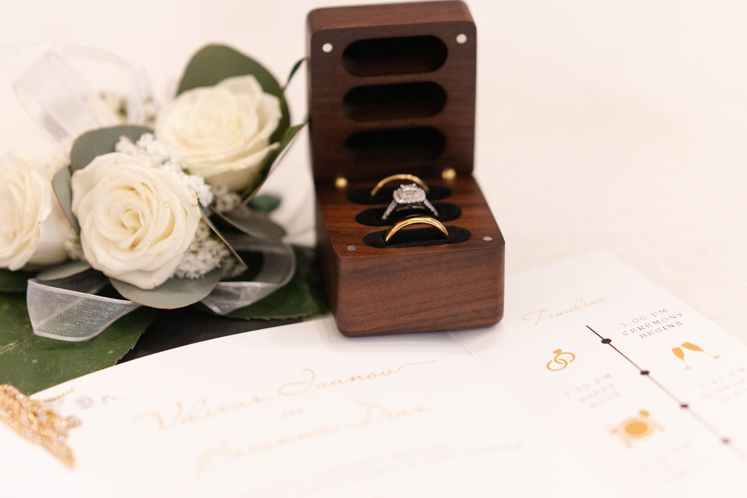 Wedding rings displayed in a wooden box next to white roses and papers on a light surface.