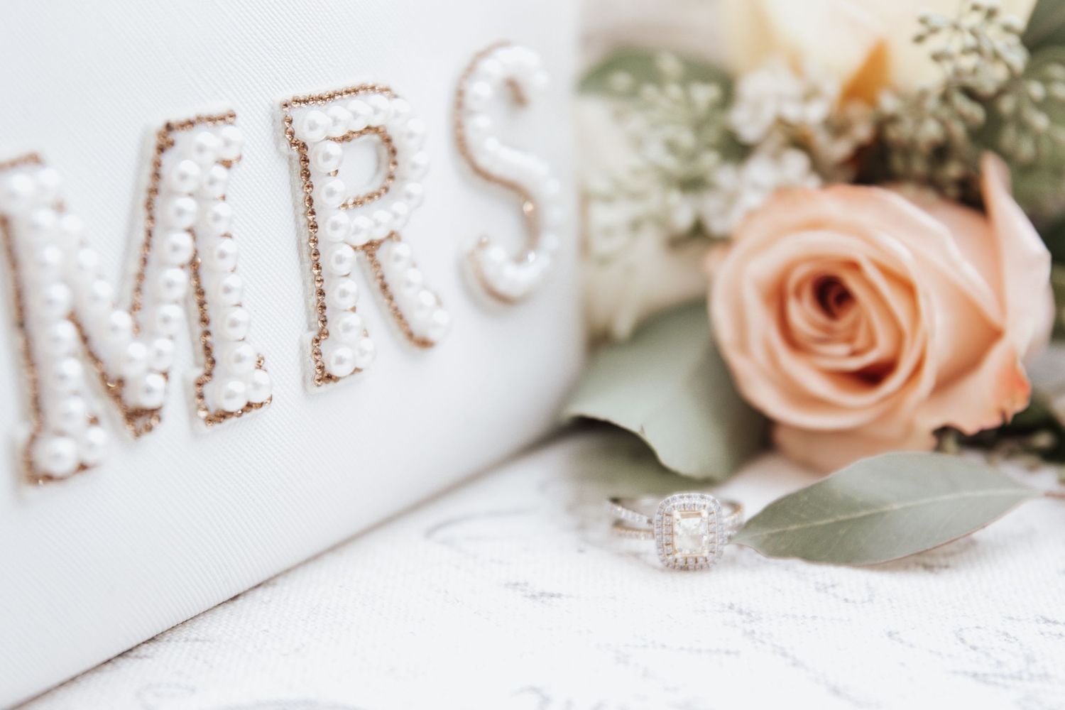 Wedding details with 'MRS' text decoration, a peach rose, and engagement ring on white surface.