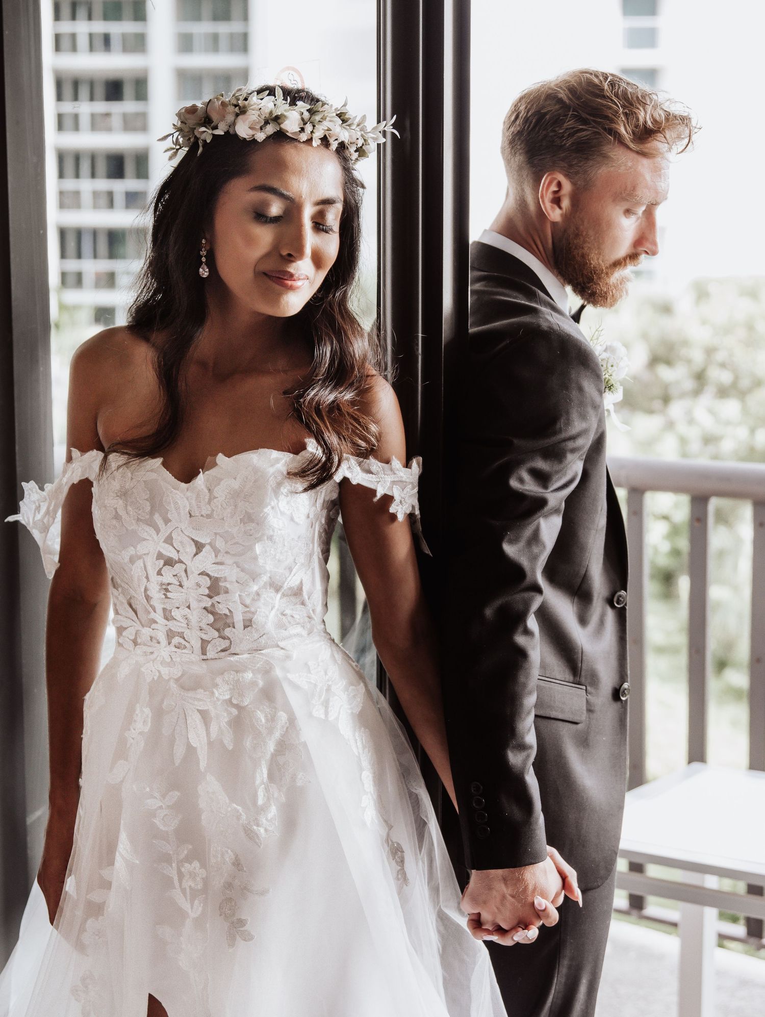 A romantic first look moment between bride in lace gown and groom in gray suit by glass windows.