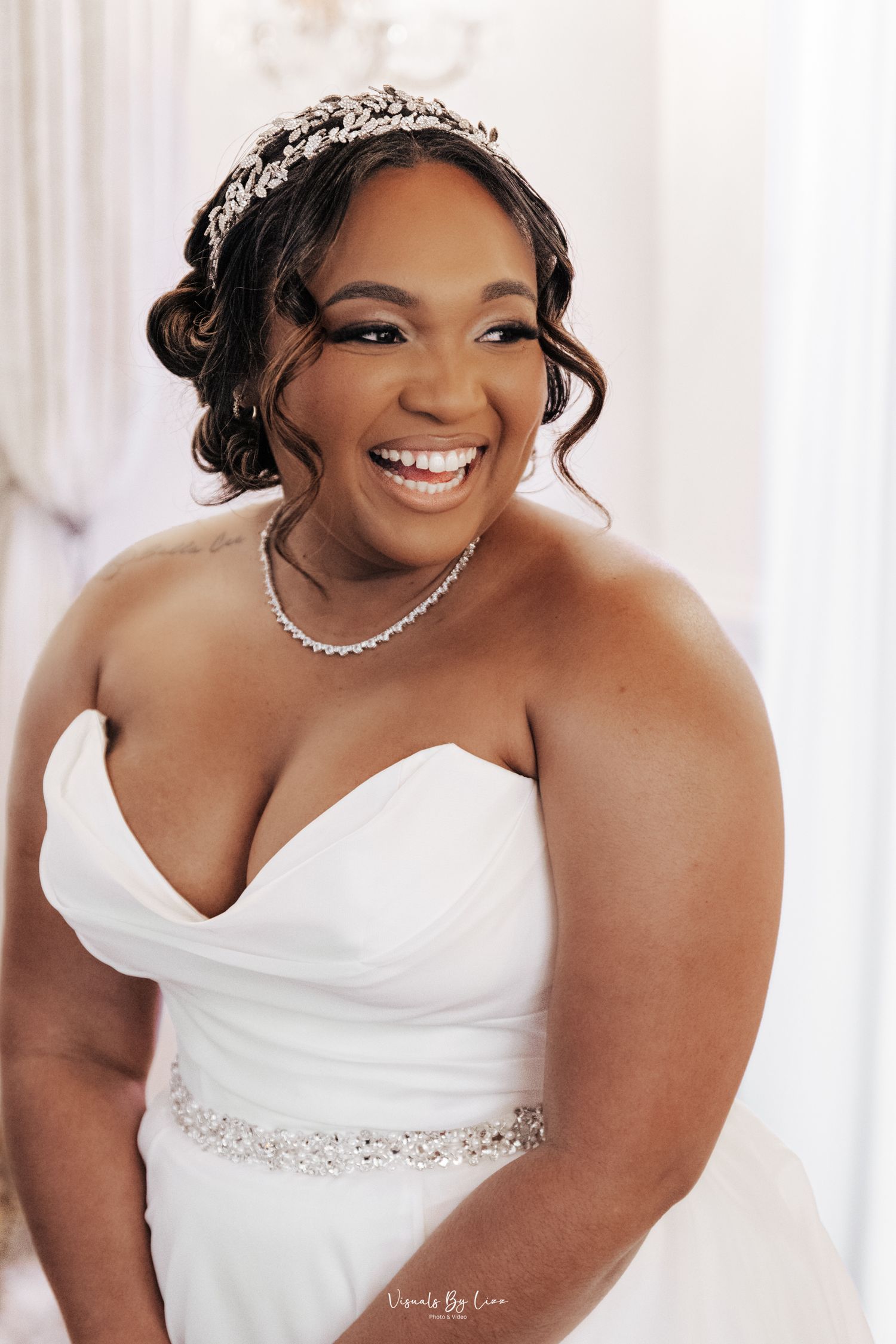 Bride wearing strapless white wedding gown with crystal belt, tiara and necklace smiling radiantly in natural light.
