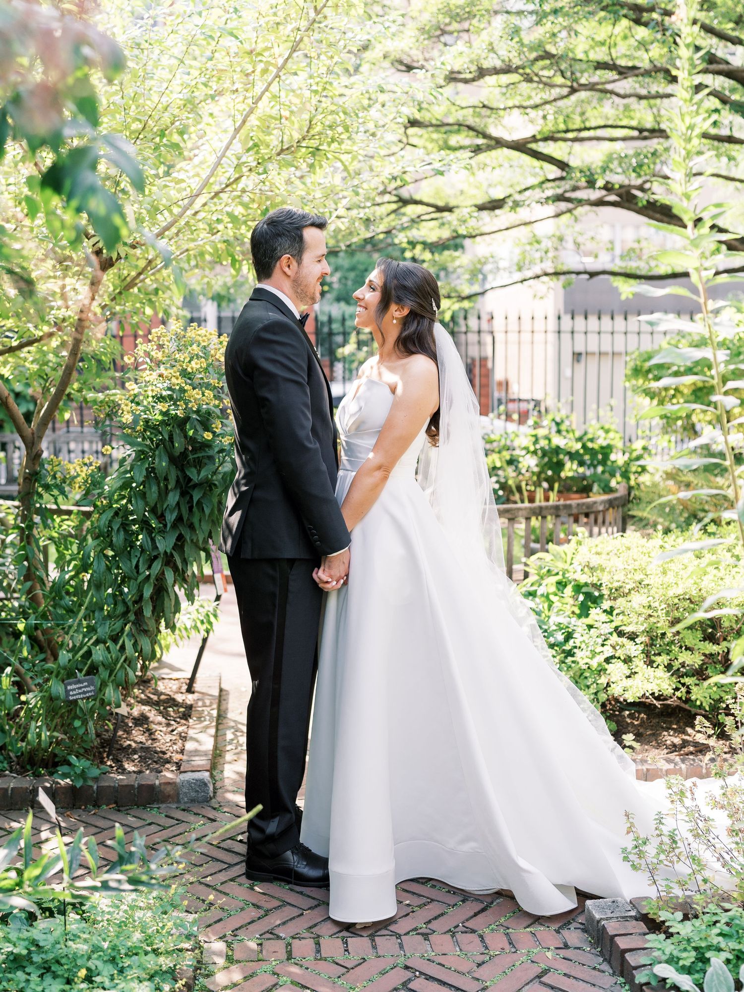 Bride and groom hold hands in a lush garden, exchanging loving glances in wedding attire.