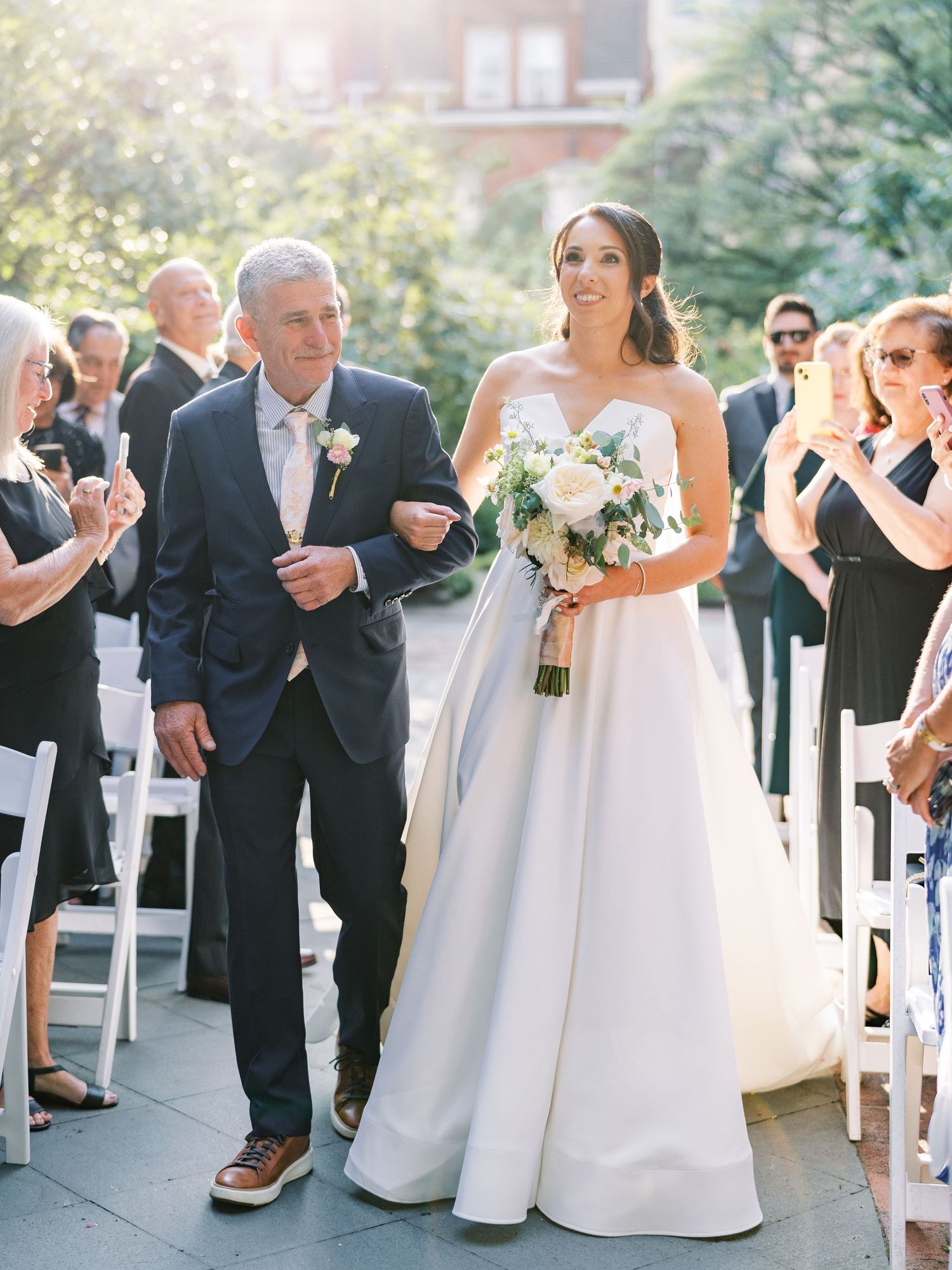 Bride in white strapless gown walks down outdoor aisle with father, holding floral bouquet, guests applauding in sunlight.