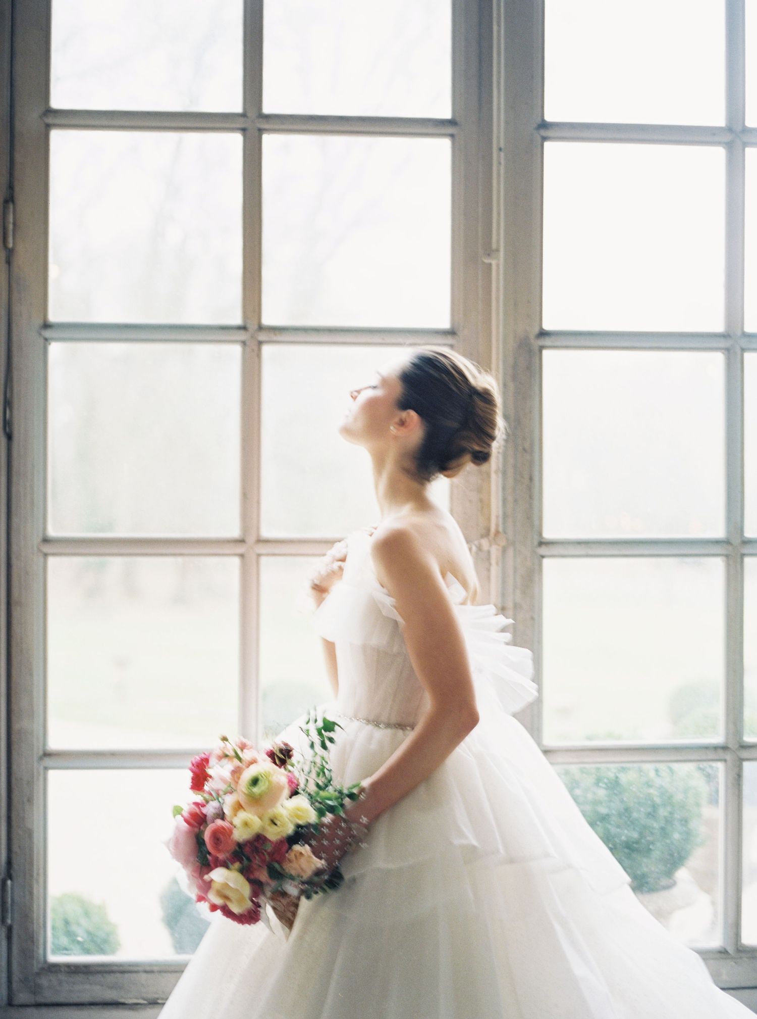 Bride in a white tulle ball gown holding a colorful bouquet, standing by a large window in soft natural light. Film Wedding Photography