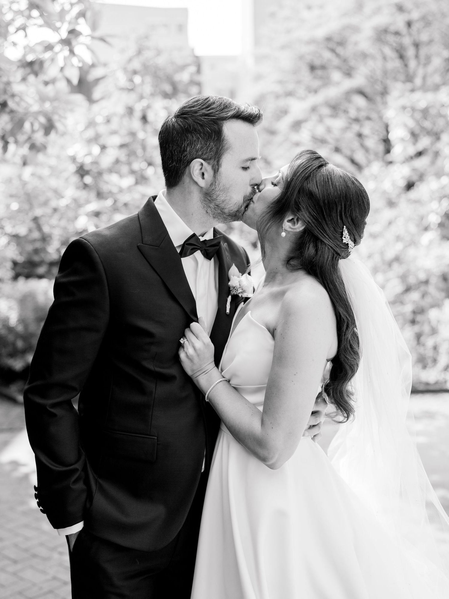 Black and white photo of a bride and groom sharing a romantic kiss on their wedding day outdoors.