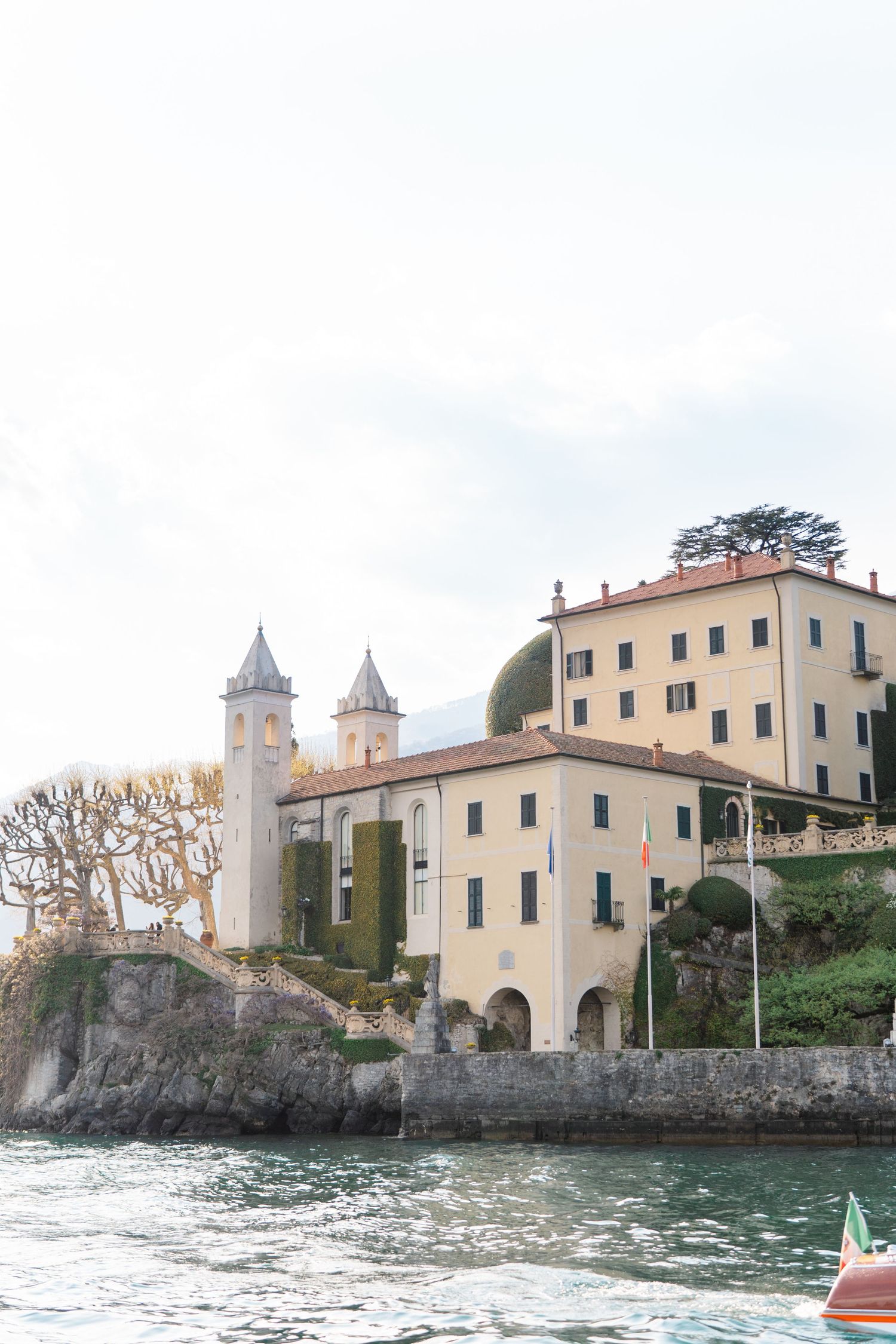 Historic Italian villa with twin towers on Lake Como shoreline, surrounded by lush greenery and stone walls under overcast skies.