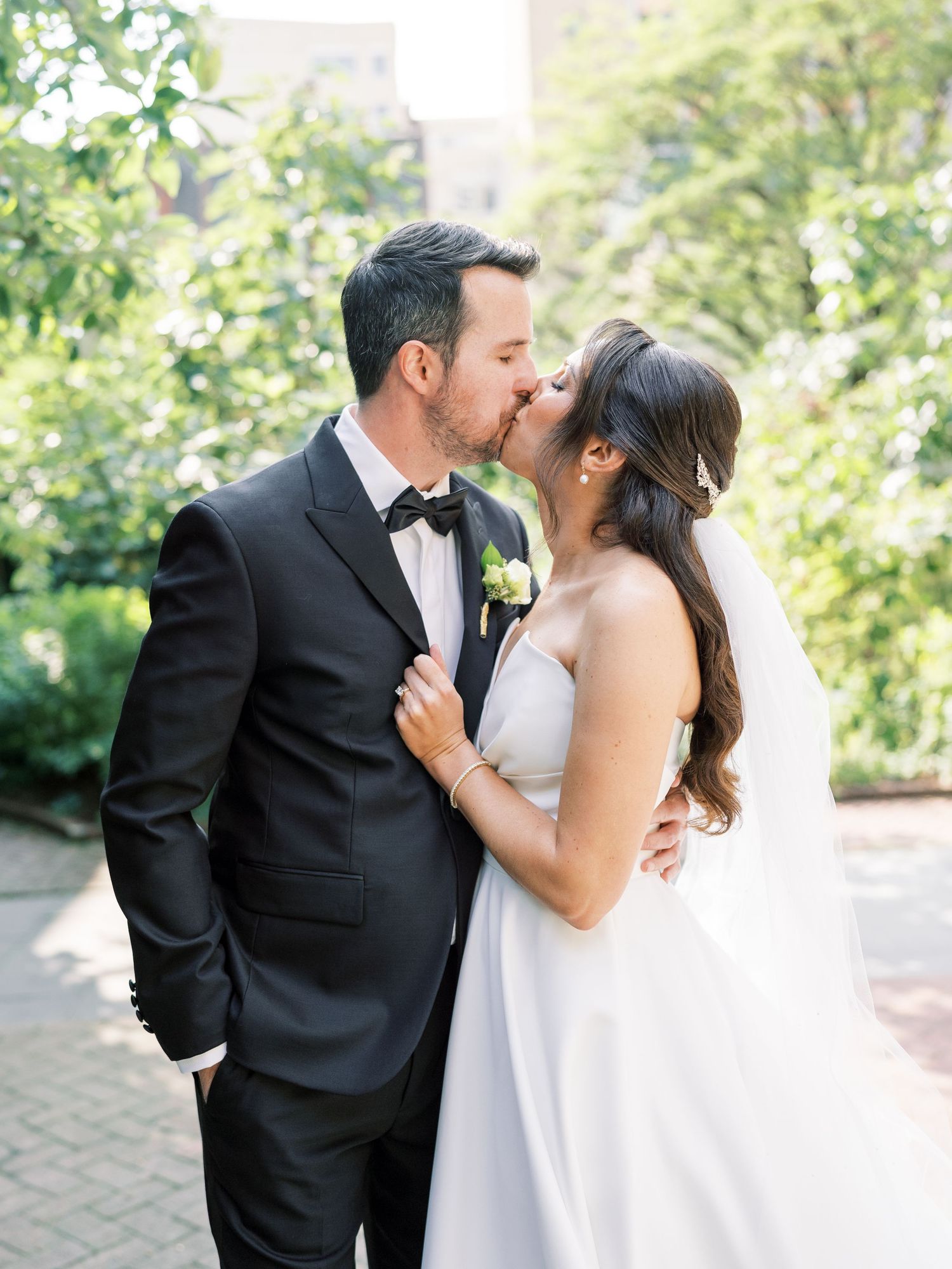 A bride and groom share a romantic kiss outdoors, she in a white gown with veil, he in a black tuxedo with bow tie.