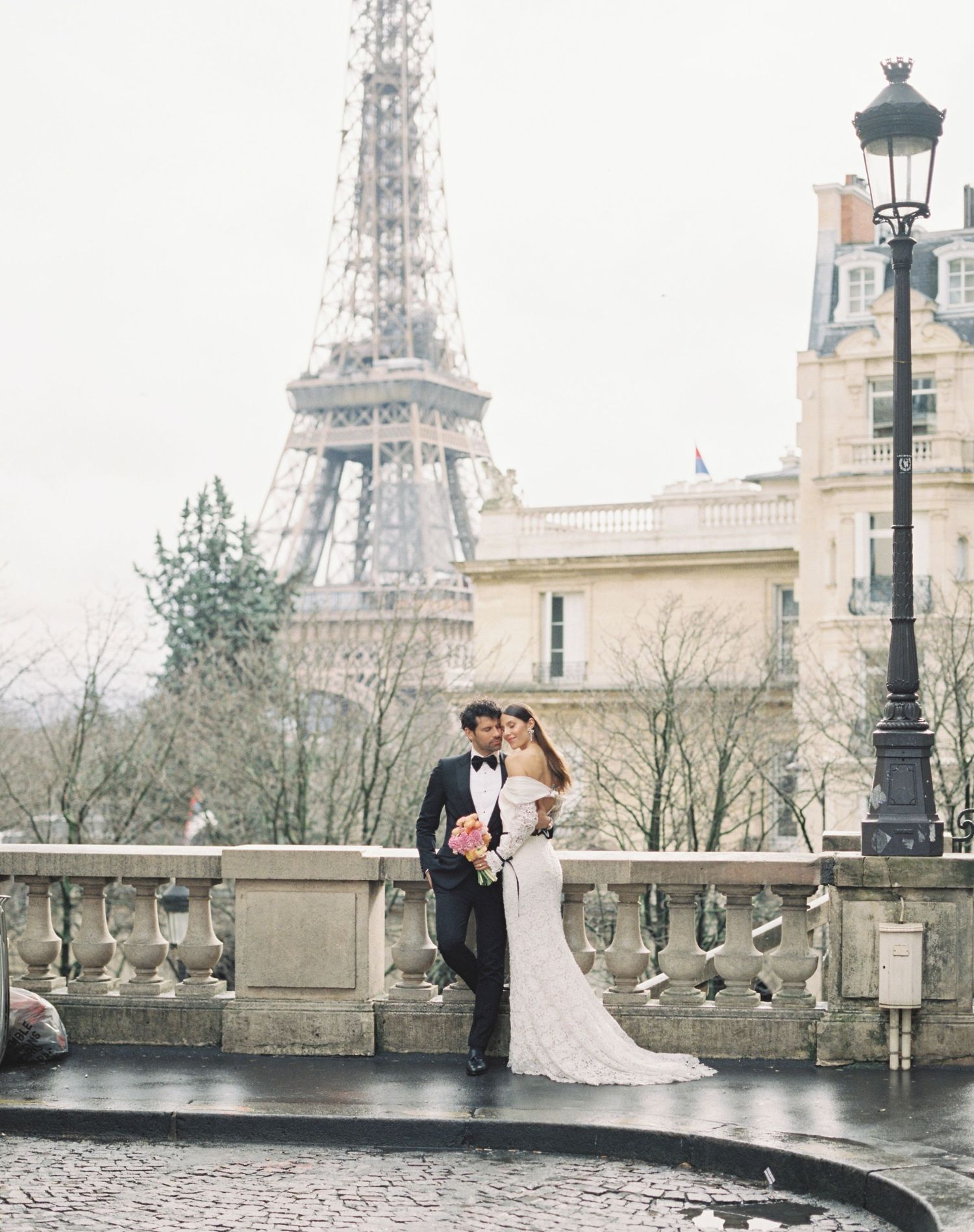 Couple in wedding attire embracing near the Eiffel Tower in Paris on a misty day.