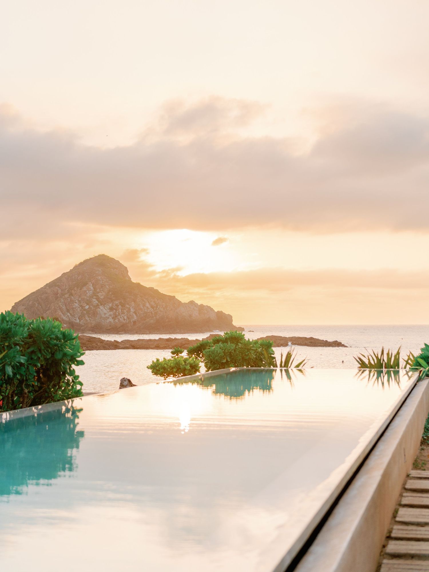 Infinity pool overlooking a rocky island and golden sunset over the ocean, surrounded by lush tropical greenery. Mexico Wedding