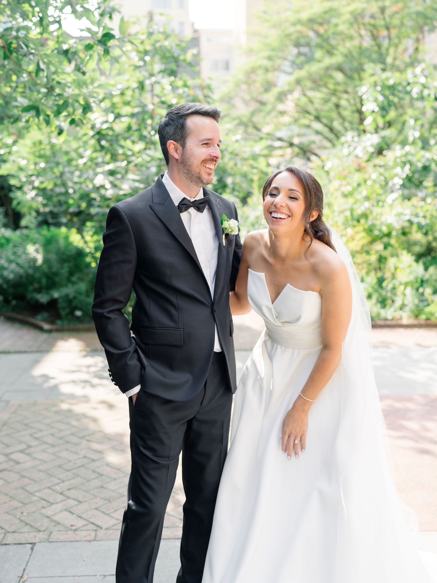 A smiling bride in a strapless white gown and groom in a black tuxedo pose together outdoors on their wedding day.