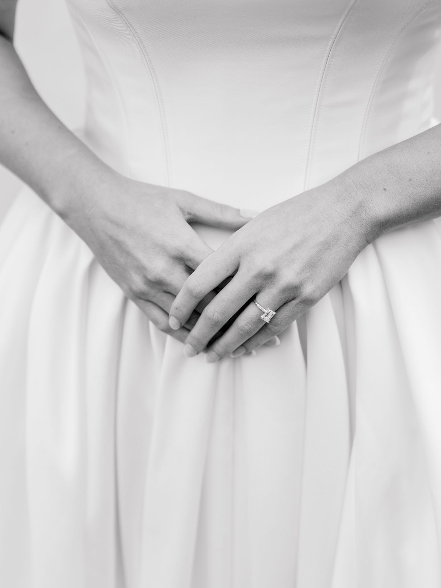 Black and white photo of a bride in a white gown clasping her hands. Classic, Elegant emerald-cut diamond engagement ring.