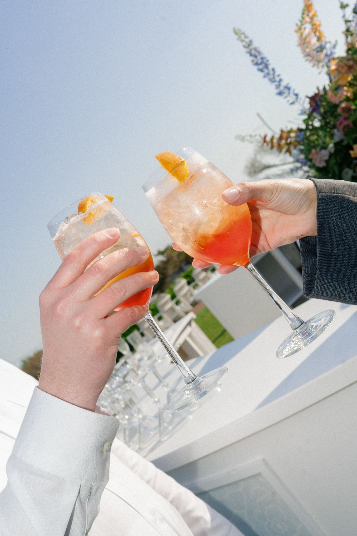 Two hands clinking cocktail glasses with orange garnishes at an outdoor wedding reception on a sunny day. Cape May New Jersey Coastal.