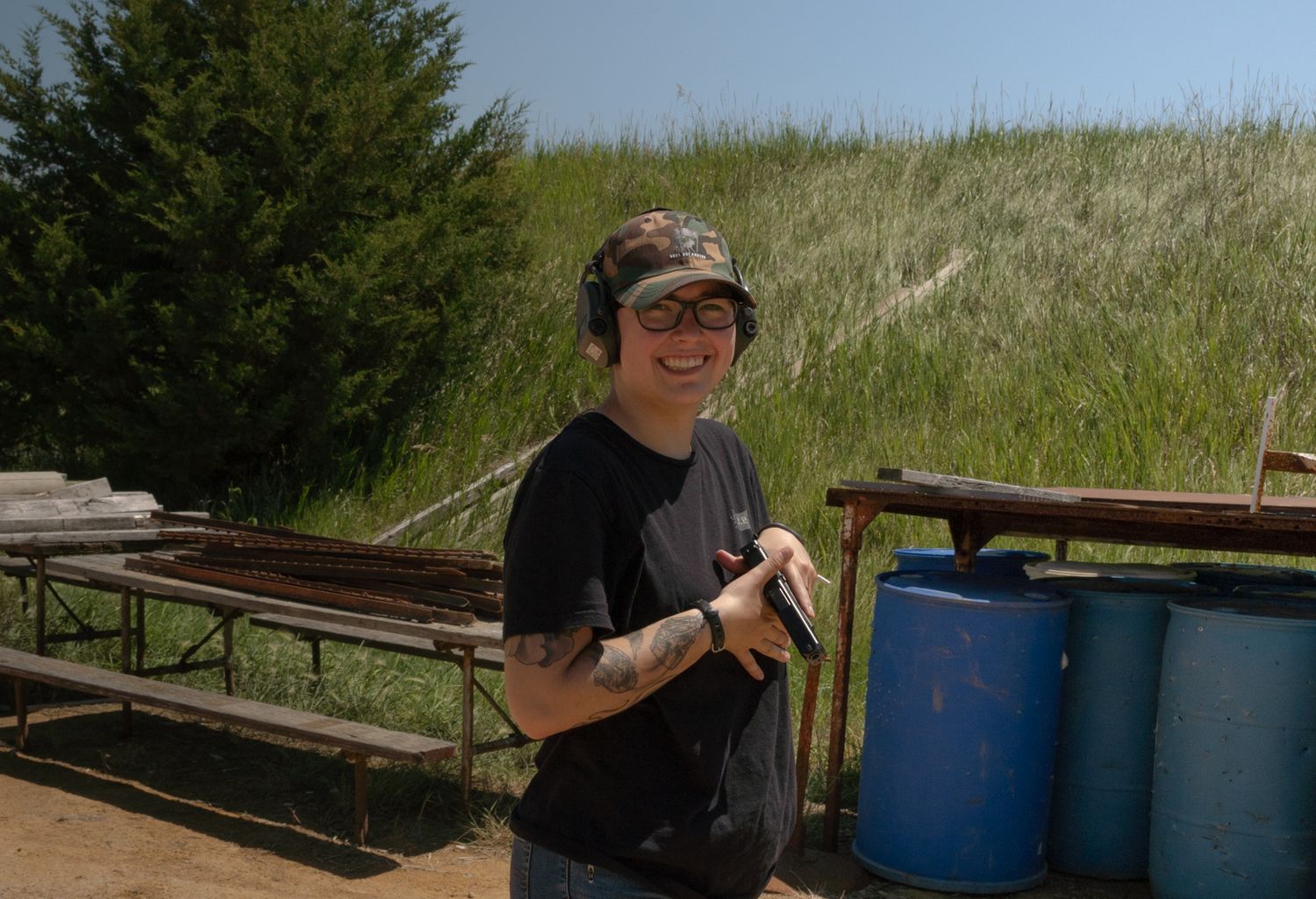 Someone wearing sunglasses and a black t-shirt stands next to blue barrels in an outdoor setting.