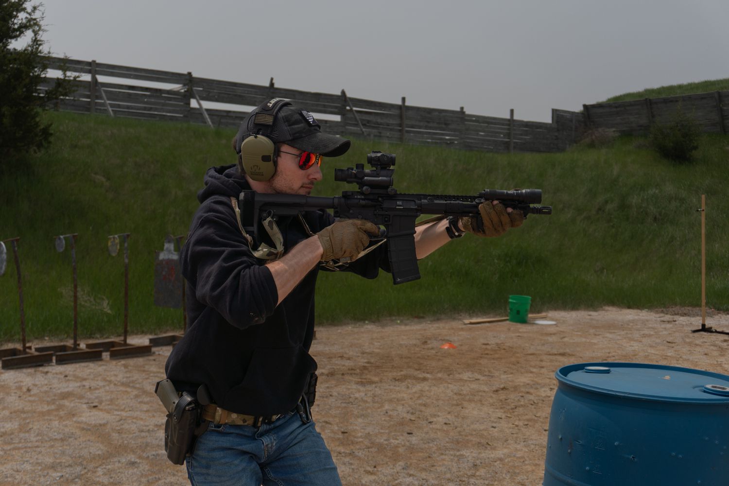 A shooter takes aim with a rifle at an outdoor target range with blue barrels and equipment visible in the background.