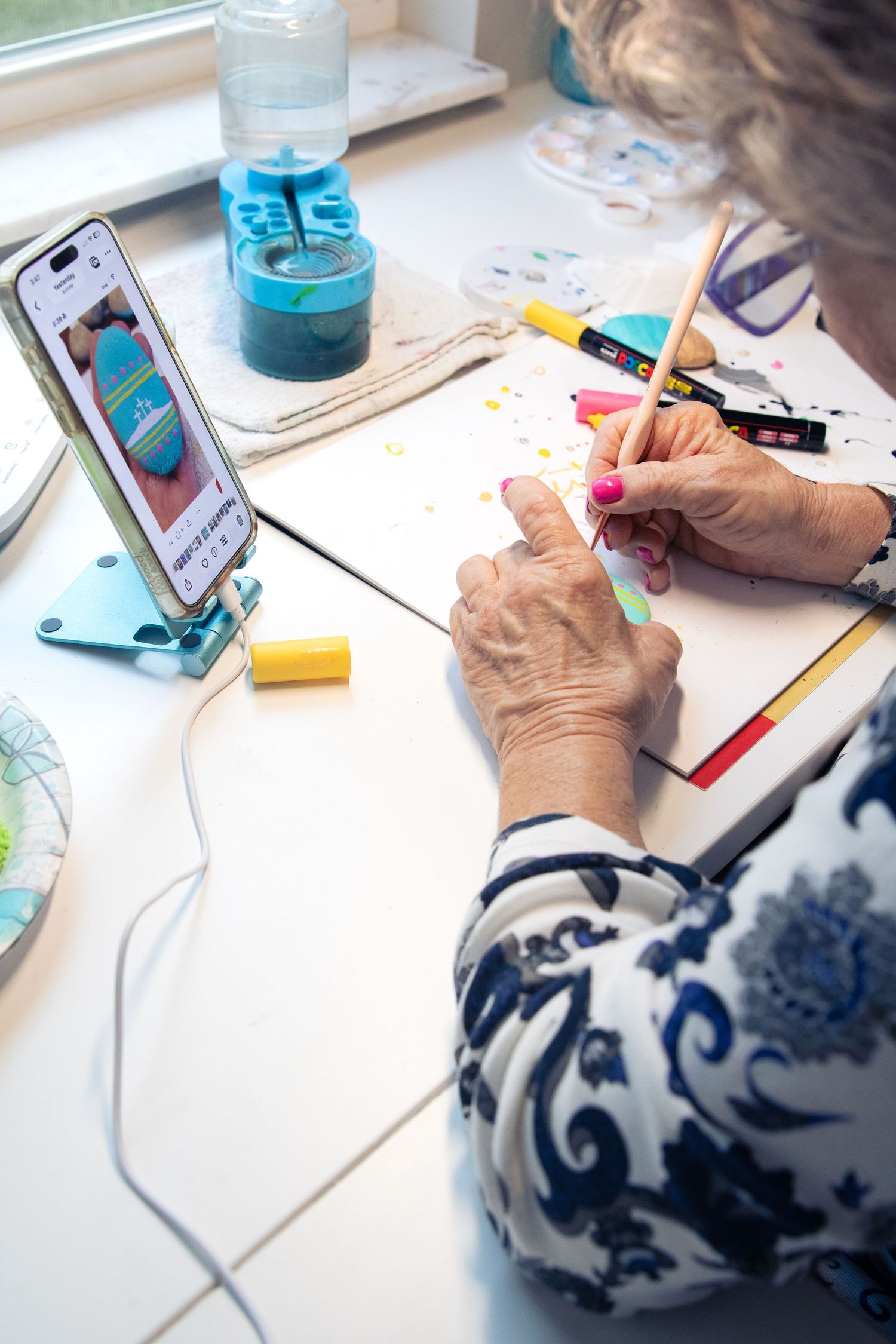branding session featuring woman painting artwork at desk using pencil while referencing tutorial on smartphone.