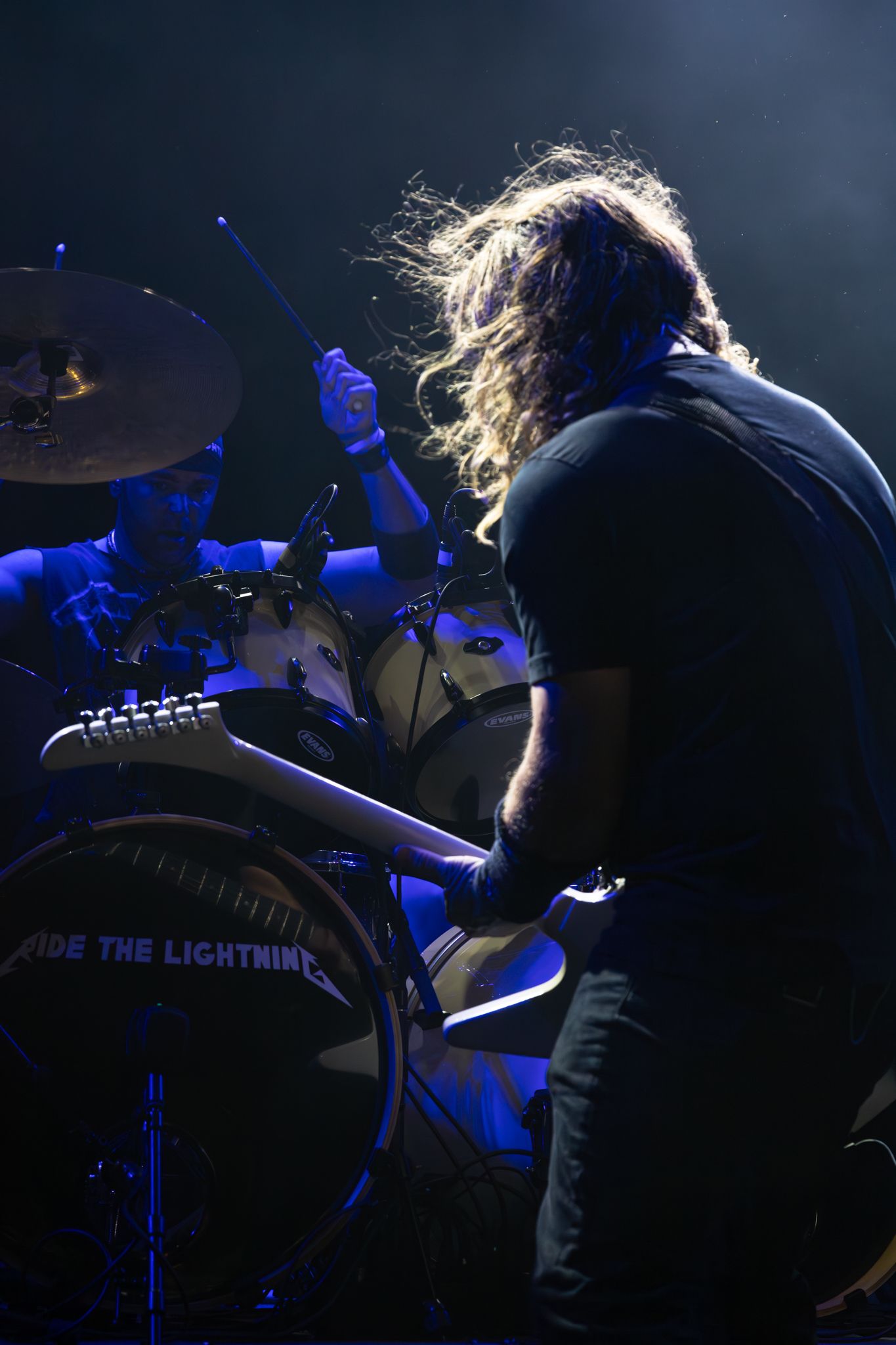 Long-haired guitarist performs on stage with drummer behind a kick drum reading Ride The Lightning in blue concert lighting.