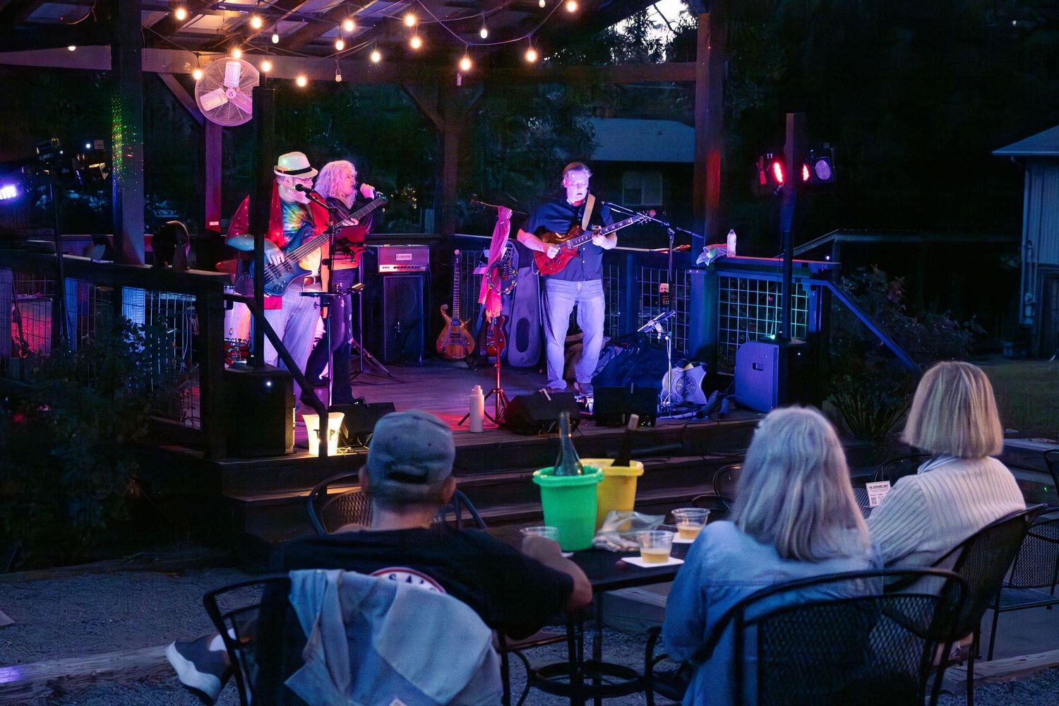 Outdoor evening concert with band performing on a lit stage while audience sits at patio tables in Monroe, NC.