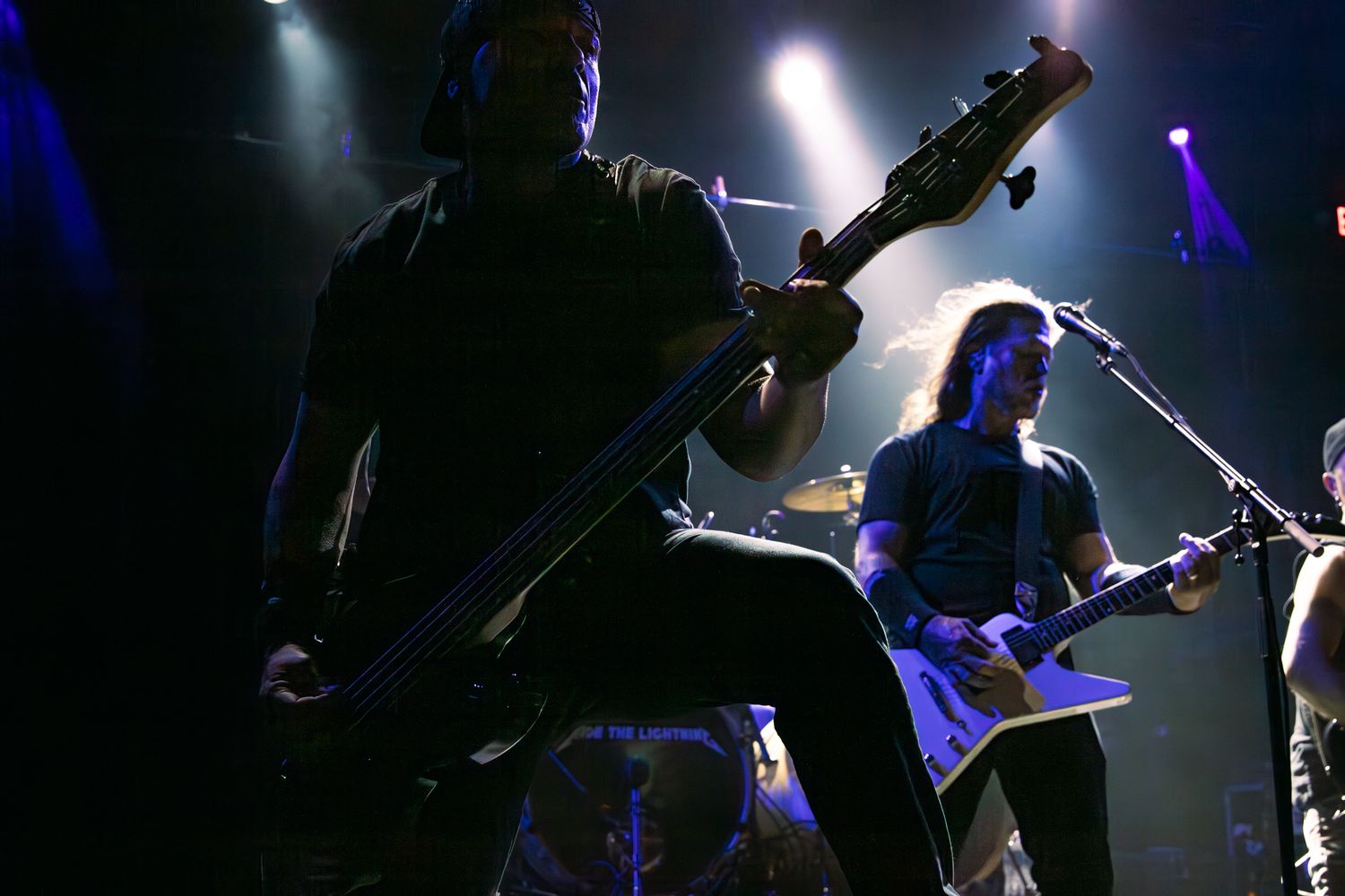 Two guitarists performing on a darkened concert stage with dramatic blue and purple lighting.