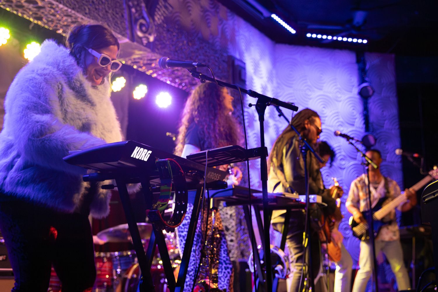 Female keyboardist in white fluffy jacket and sunglasses performs alongside a band on a lit stage in Charlotte, NC.