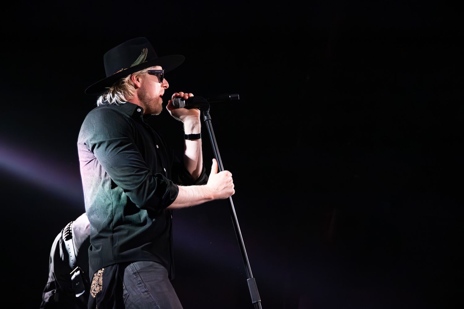 Male singer in black hat performing passionately into microphone under dramatic spotlight on stage in Charlotte, NC.