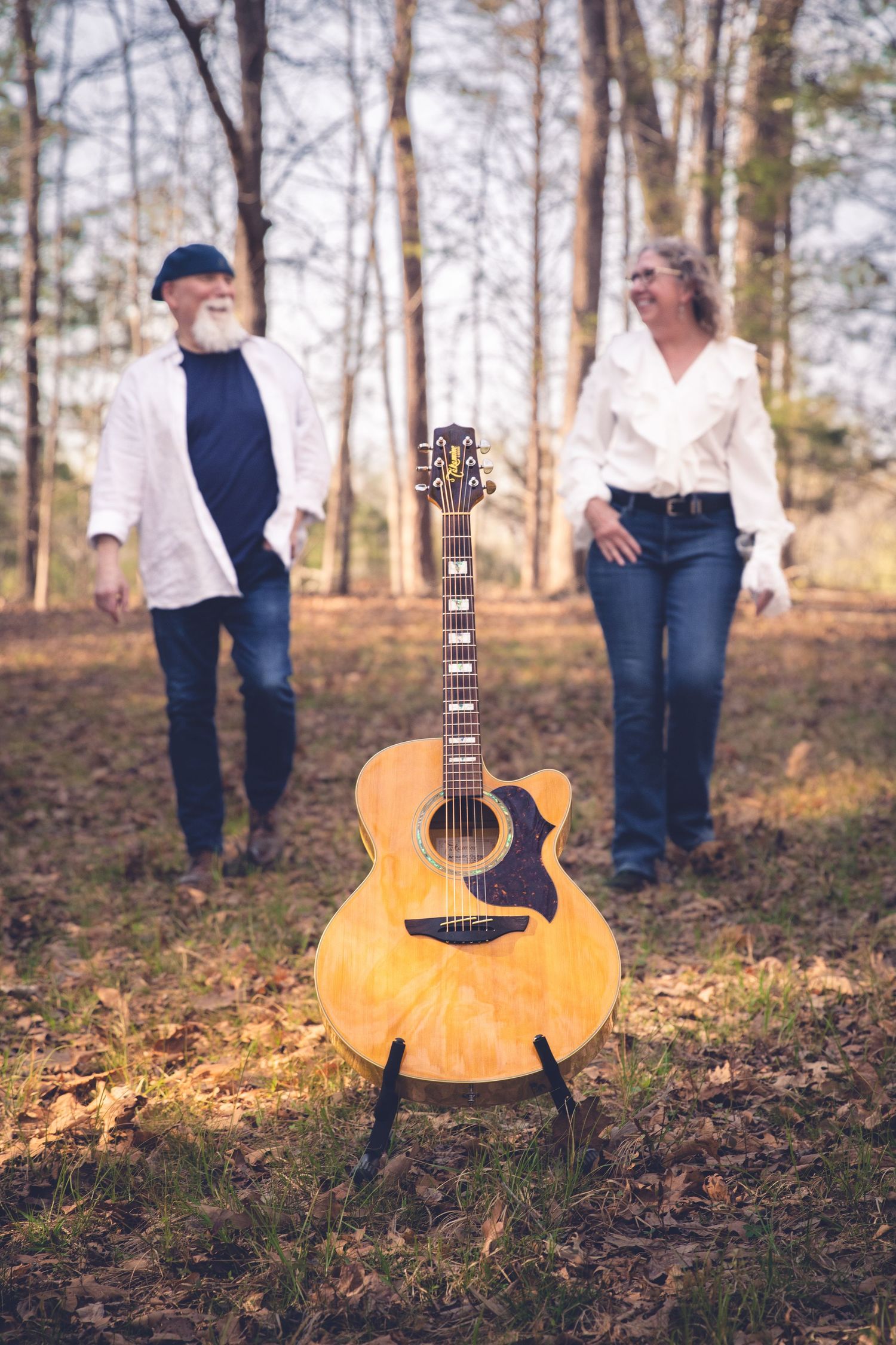 promotional band photo featuring acoustic guitar on stand in forest with two adults in white shirts and jeans standing in background.