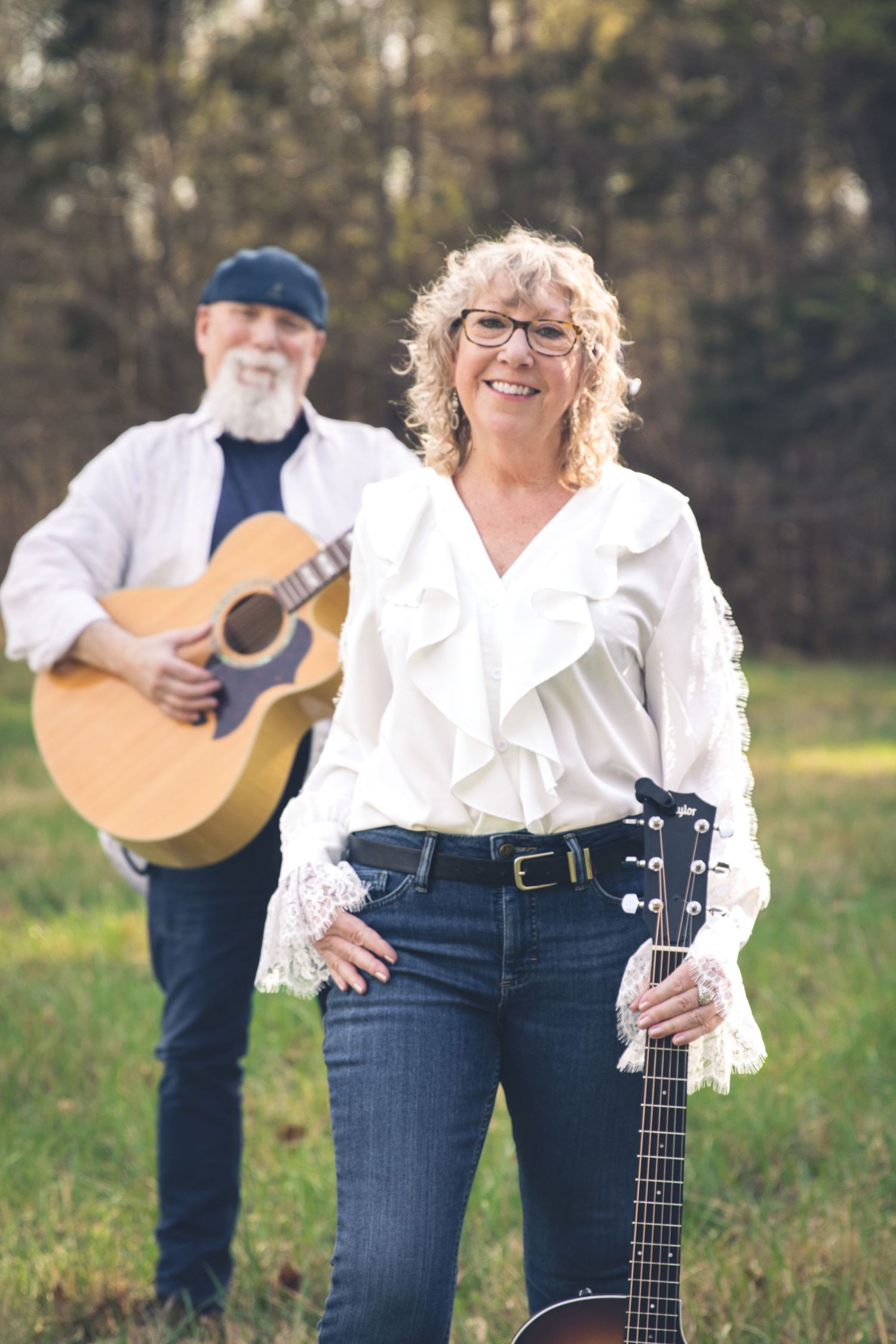 Promotional band photo featuring smiling woman with glasses holds a guitar outdoors while a bearded man plays acoustic guitar behind her.