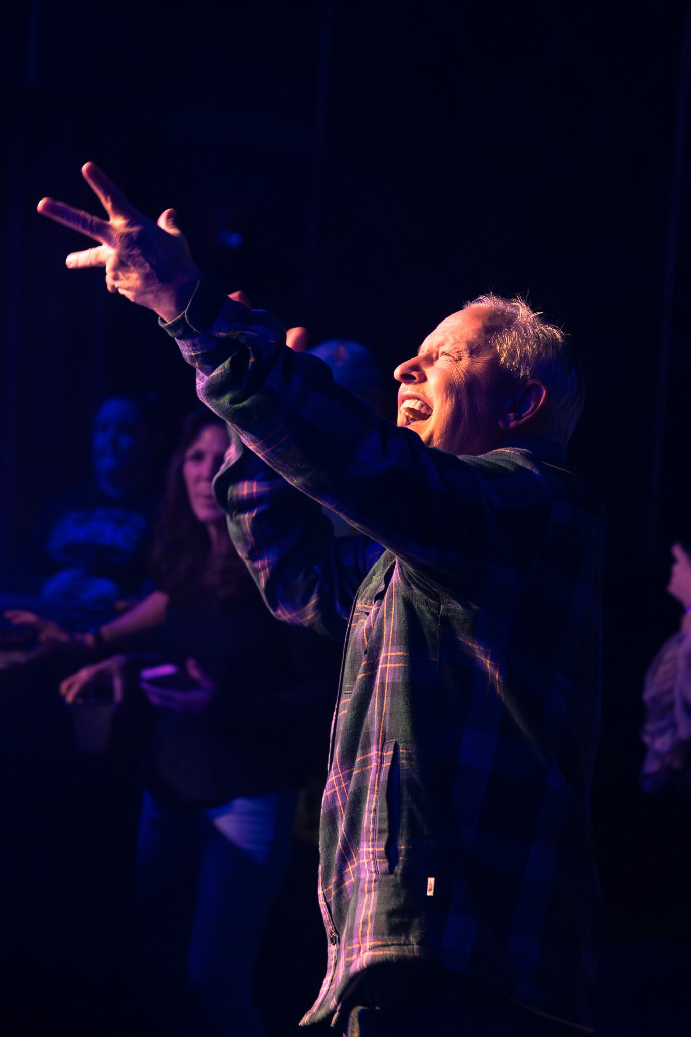 Older man passionately singing on stage with arms raised under blue spotlight.