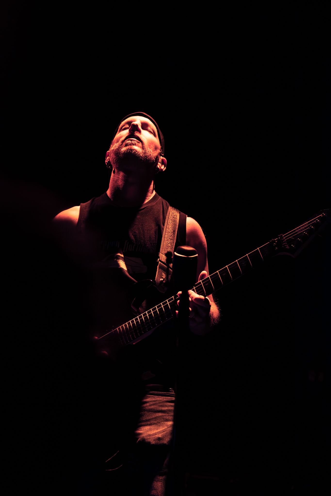 A bearded male guitarist performing on stage, dramatically lit in red light against a dark background.
