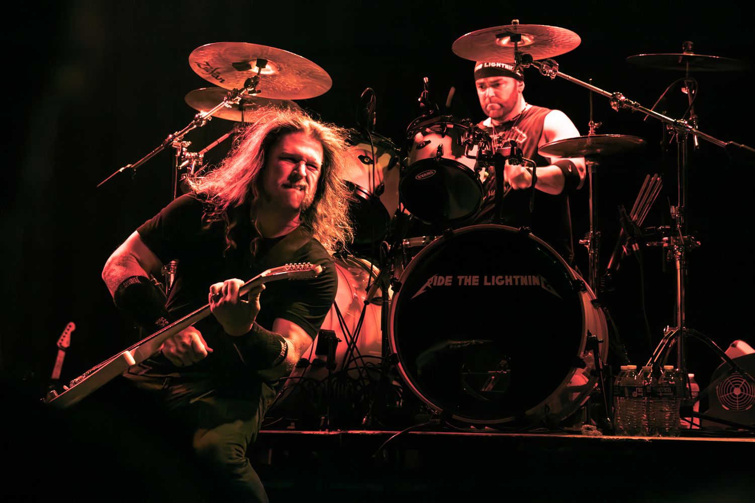 Two heavy metal musicians perform live, a long-haired guitarist and a drummer on stage under dramatic red lighting.