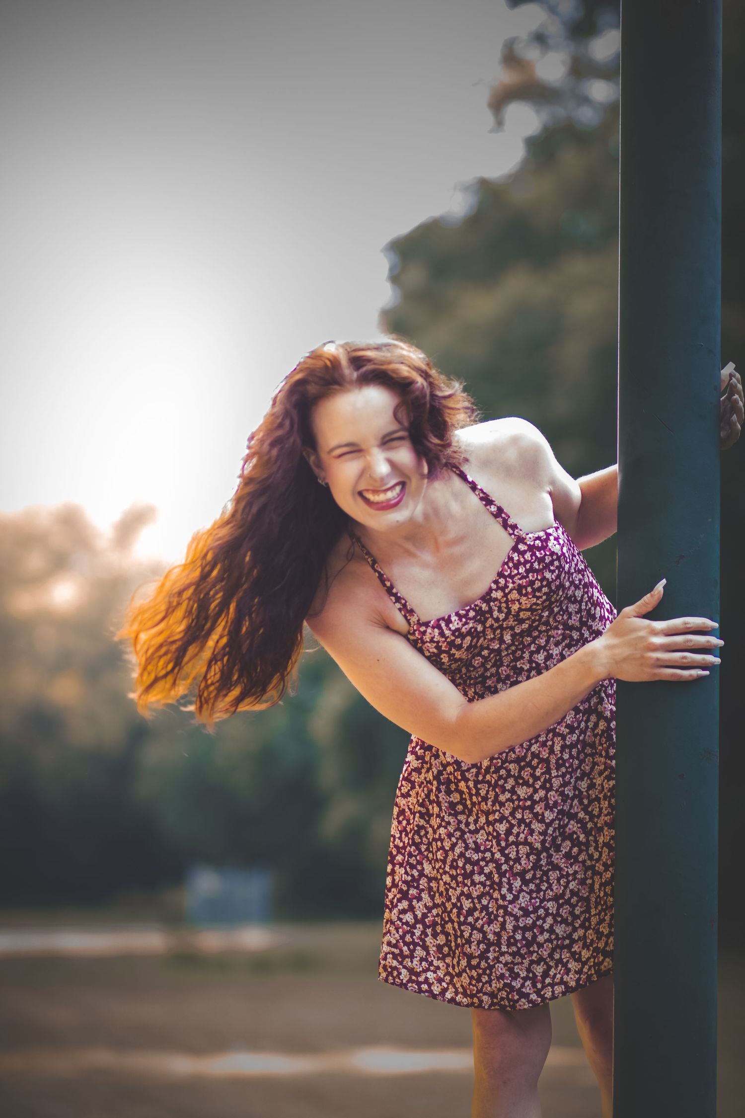 Cheerful brunette woman in floral sundress twirling around metal pole at sunset outdoors.