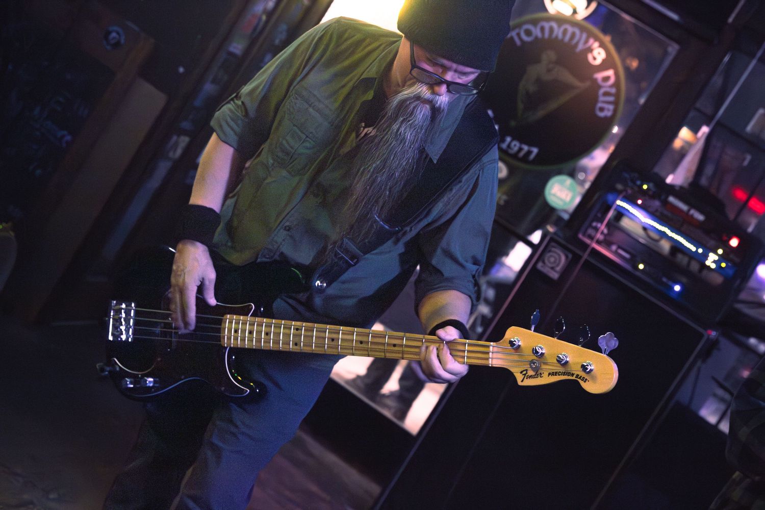 Bass guitarist wearing cap and green shirt playing black Fender bass guitar on dark stage.