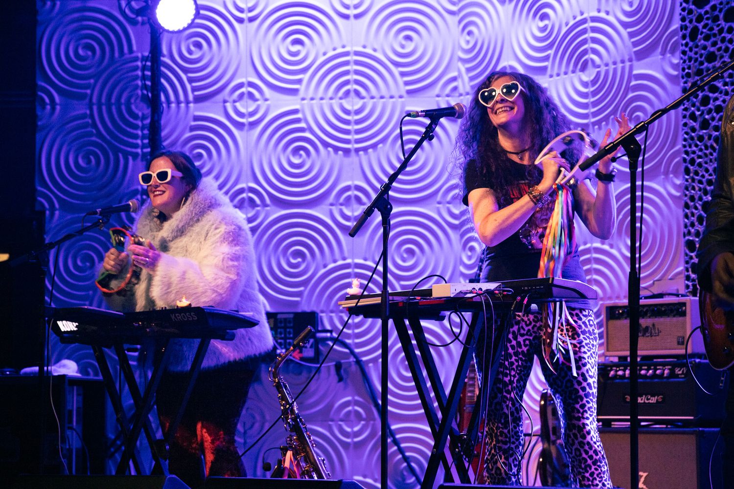 Two female musicians perform on stage with keyboards, wearing sunglasses against a purple swirl backdrop.